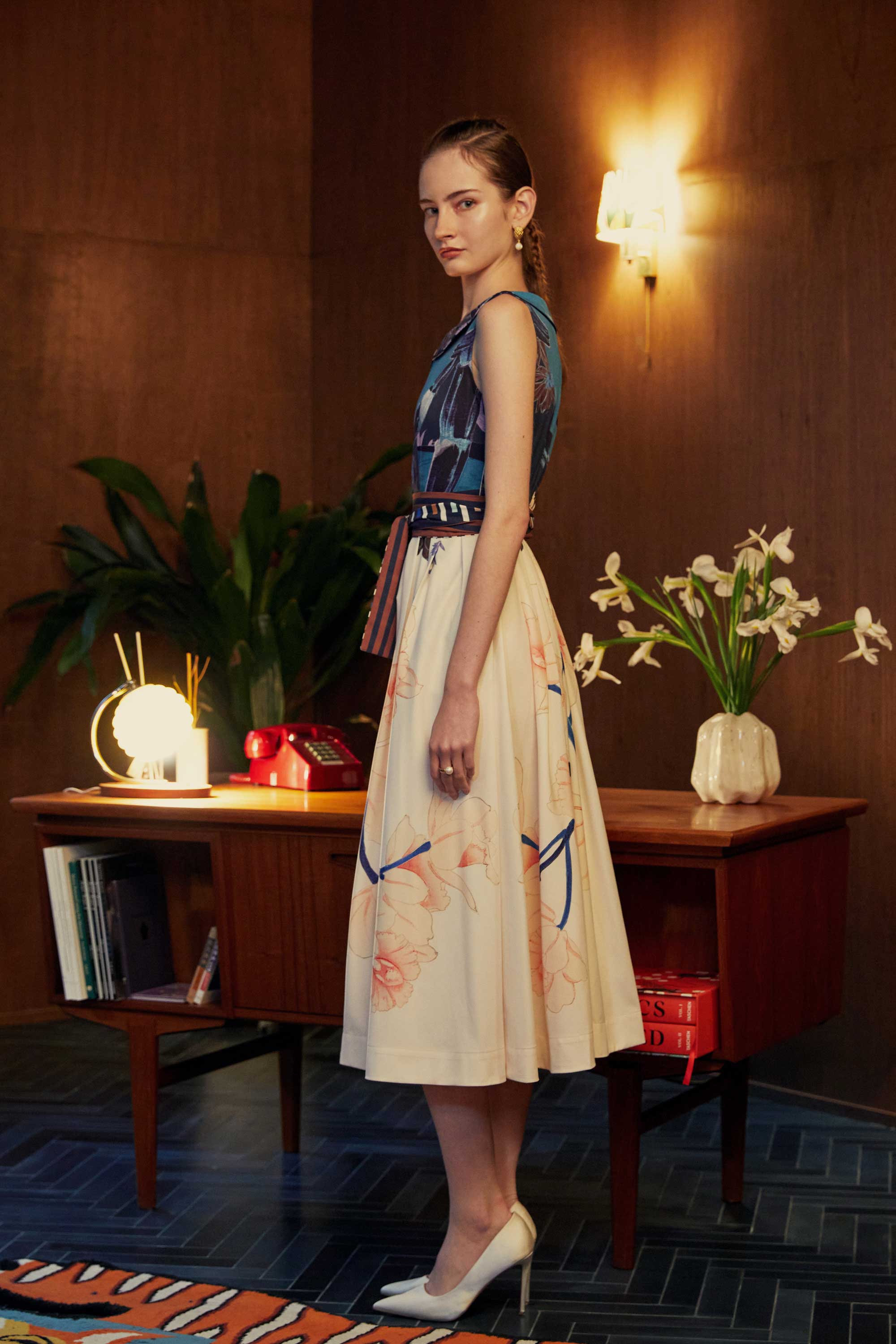 Side view of model in the BEHOLD Peter Pan Fit-and-Flare Midi Dress in Dendrobium Chrysotoxum orchid print, featuring navy sleeveless bodice with Peter Pan collar, cream flared midi skirt with orchid floral motifs, and striped waistband, styled indoors with wood-paneled backdrop.