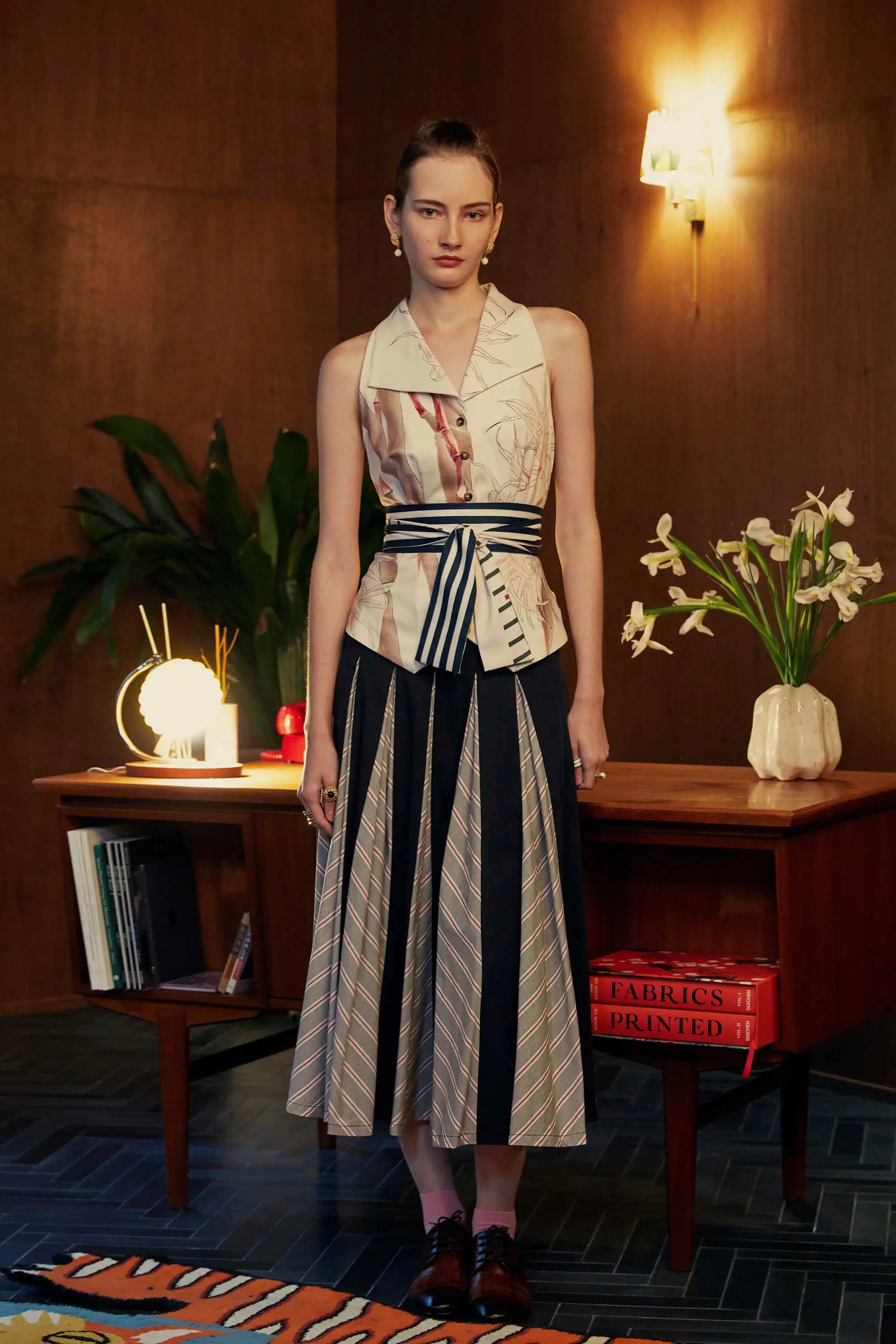 Front view of woman in a racerback vest with orchid print and striped sash, paired with a pleated godet skirt featuring bold stripe inserts, styled with black leather shoes in a mid-century styled room.