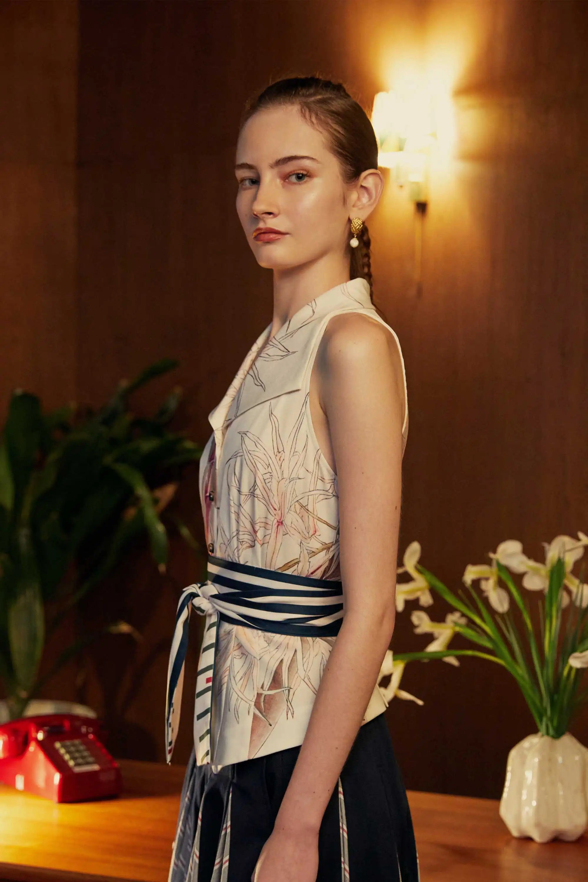Side view of woman wearing a structured sleeveless vest with orchid motifs and a striped sash belt, paired with a pleated godet skirt in a warmly lit room with floral decor and a vintage red phone.