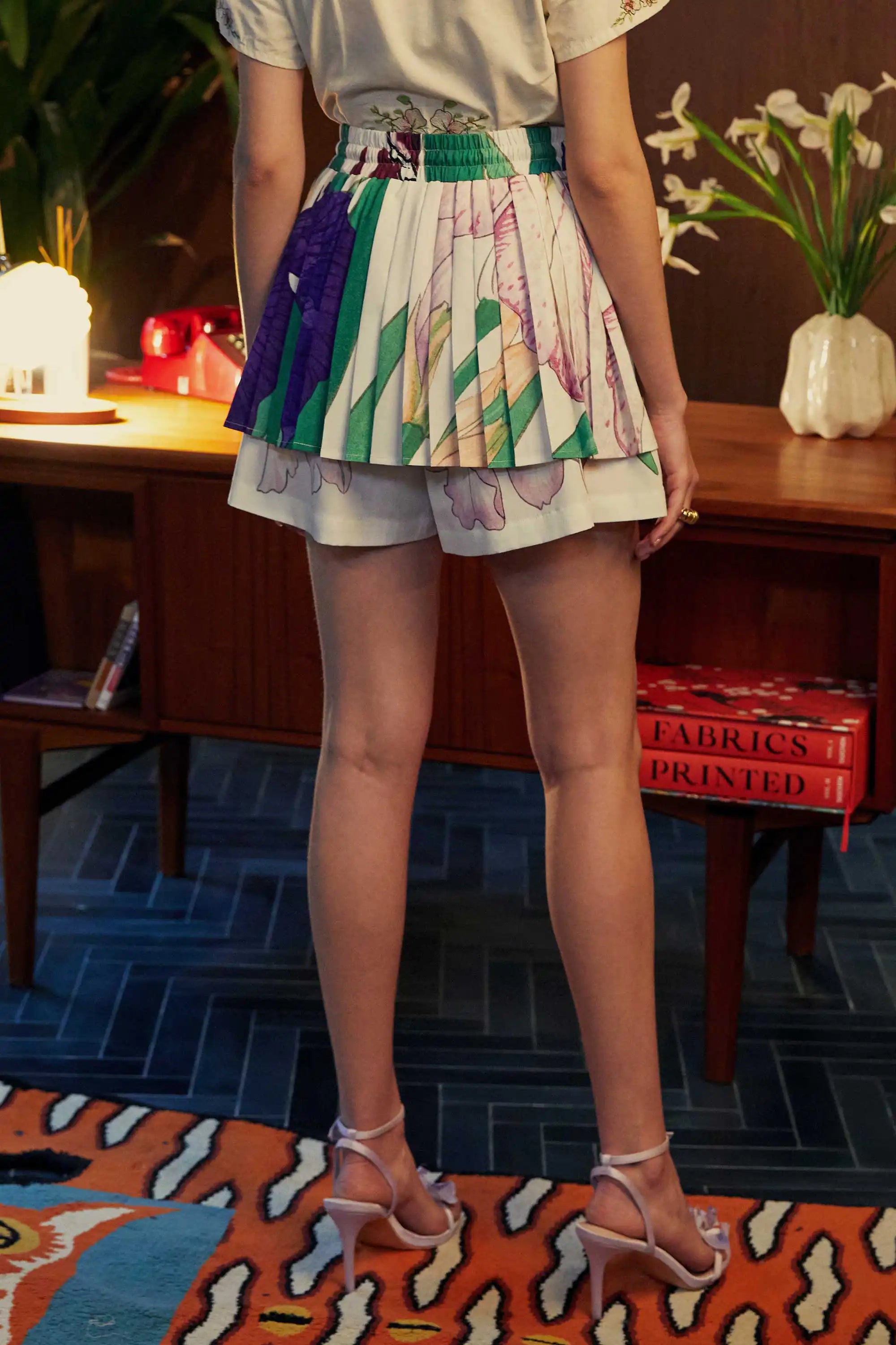 Back view of Iris-print pleated twirl skorts with built-in shorts, styled indoors with mid heels and a floral embroidered top, against a vintage wooden desk and fresh lilies.