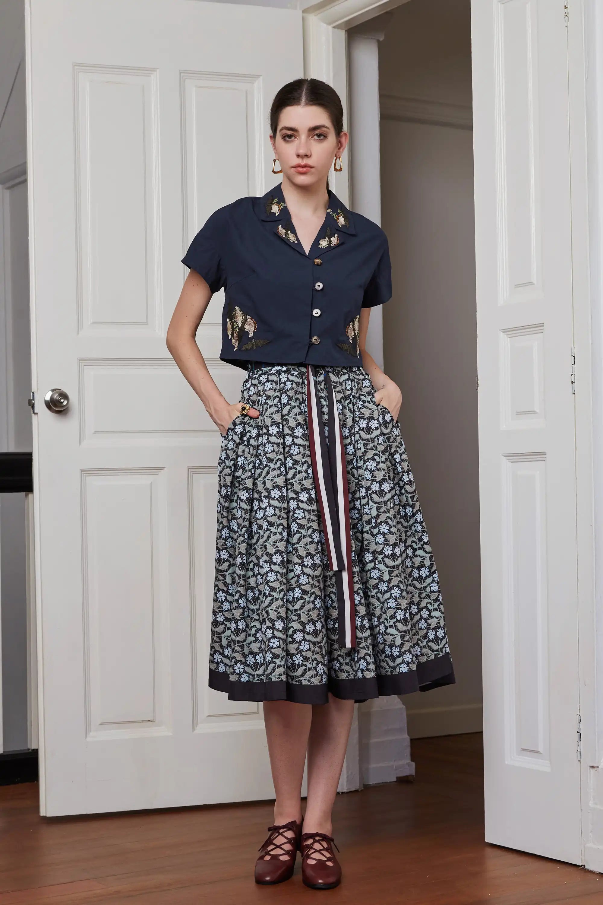 Full front view of wide leg pleated pants styled with cropped top, highlighting high waist fit, voluminous leg shape, floral print, contrast sash, and side pockets