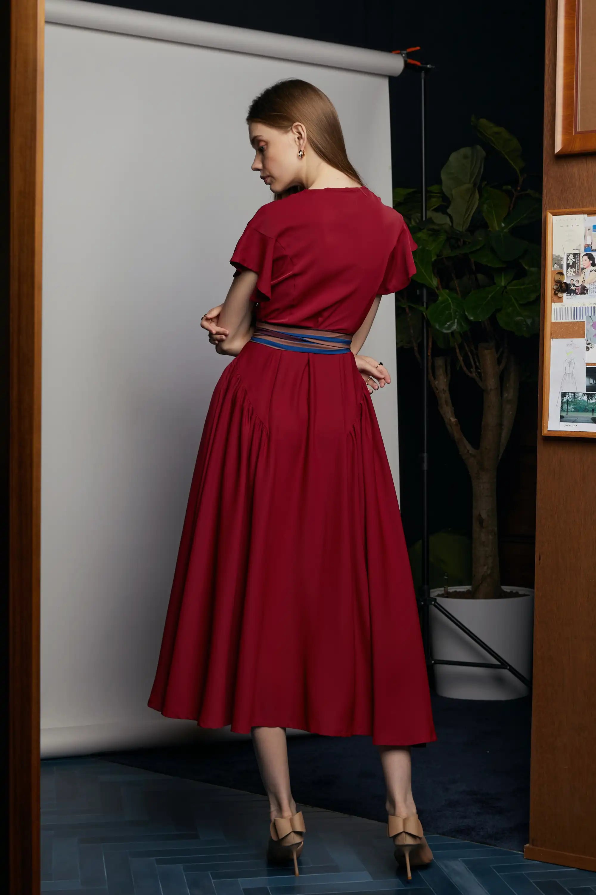 Back view of the HAVEN Bluebell Billowing Dress in deep red, showcasing the softly gathered waist and flowing maxi skirt. The look is finished with a striped sash belt featuring the Java Sparrow print. Photographed indoors against a grey backdrop with warm wood tones and a potted plant in the background.