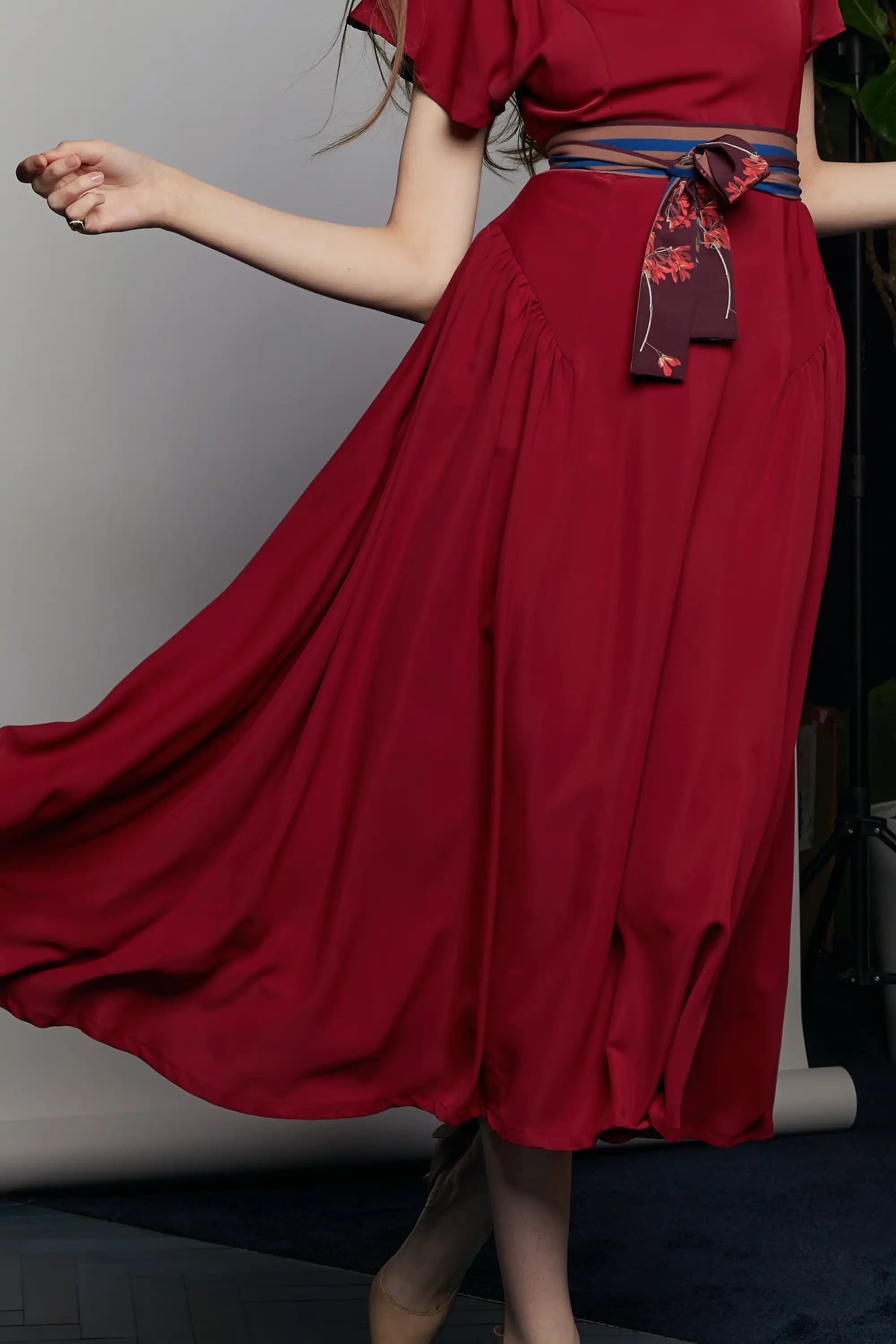 Movement shot of the HAVEN Bluebell Billowing Dress in deep red, showcasing the fluid drape of its full skirt. The waist is cinched with a striped sash belt in the Java Sparrow print, featuring a maroon accent with delicate red floral motifs. The fabric flows gracefully as the model twirls, emphasizing the dress’s light and airy construction.
