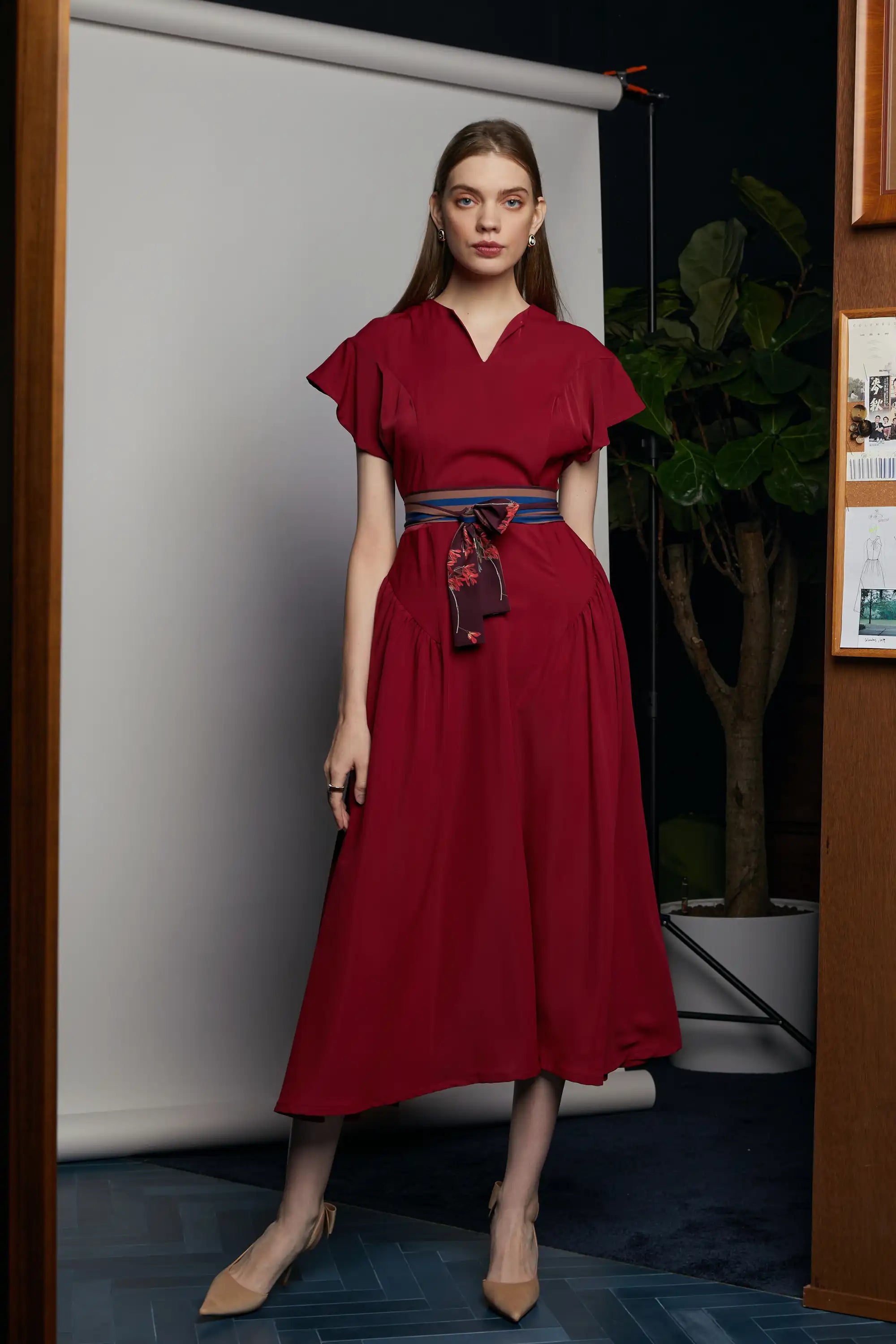 Model wearing the HAVEN Bluebell Billowing Dress in deep red with a soft V-notch neckline, flutter sleeves, and a flowing maxi skirt, cinched by a layered striped sash with a Java Sparrow bird print tied at the front, shot indoor in House of Reckless