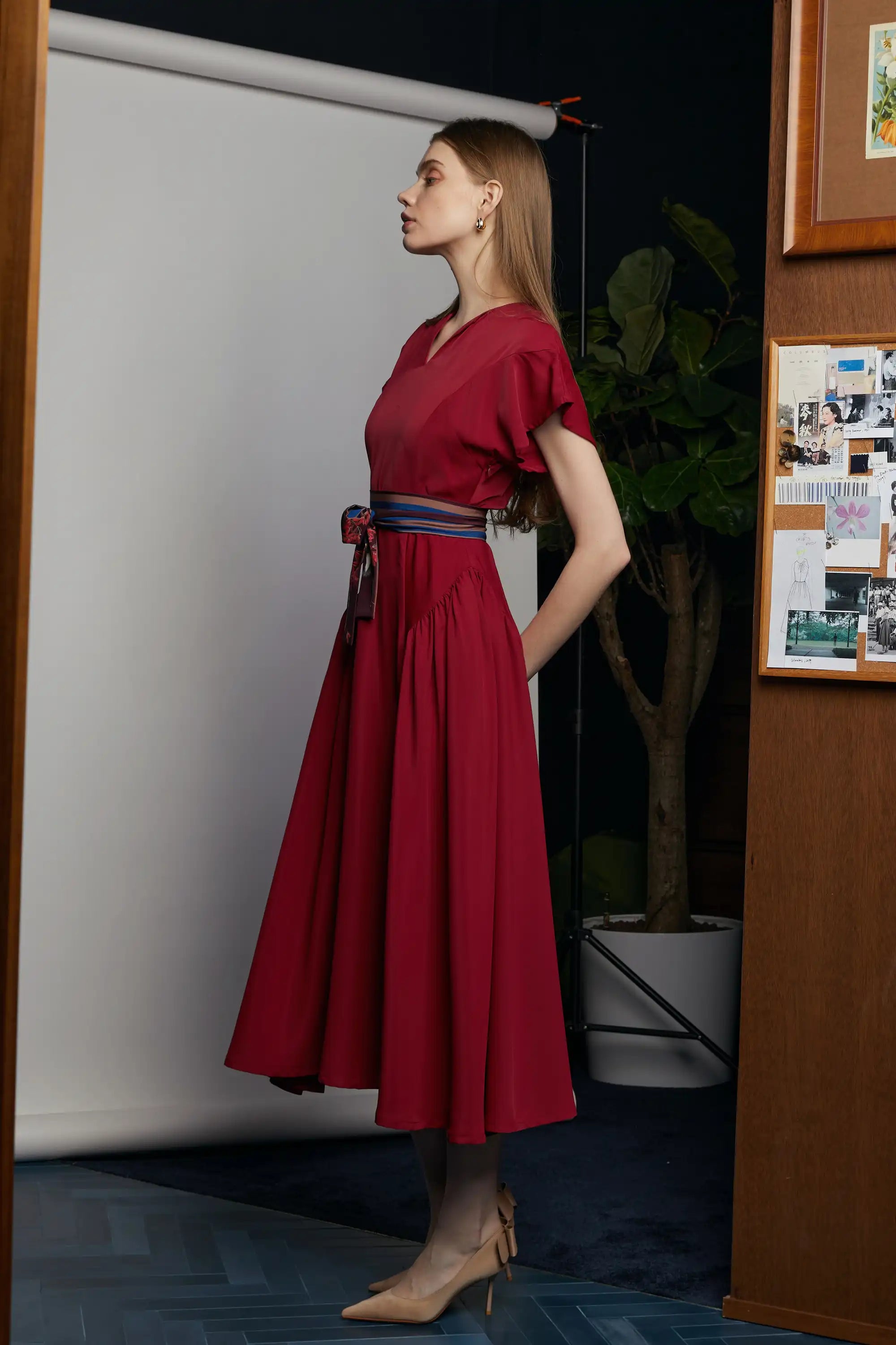 Side profile of the HAVEN Bluebell Billowing Dress in deep red, featuring a V-notch neckline, flutter sleeves, and a full billowing skirt. The waist is accentuated with a striped sash belt adorned with the Java Sparrow print, tied neatly at the side. Captured indoors with a soft backdrop, wood panel accents, and a potted plant.
