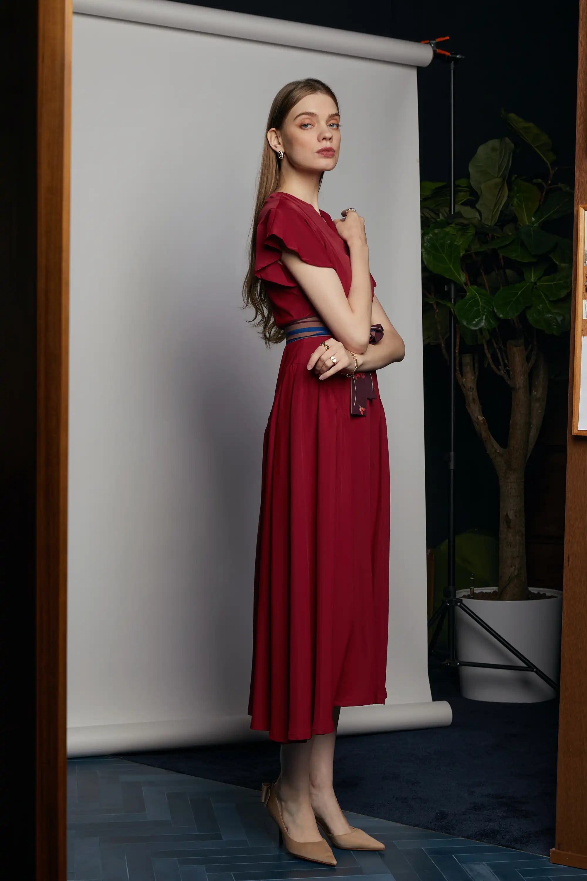 Side view of the HAVEN Bluebell Billowing Dress in deep red, highlighting its flutter sleeves, flowing maxi skirt, and striped sash belt with a Java Sparrow bird motif tied at the front. Photographed indoors against a white backdrop, with a potted fiddle-leaf fig in the background.