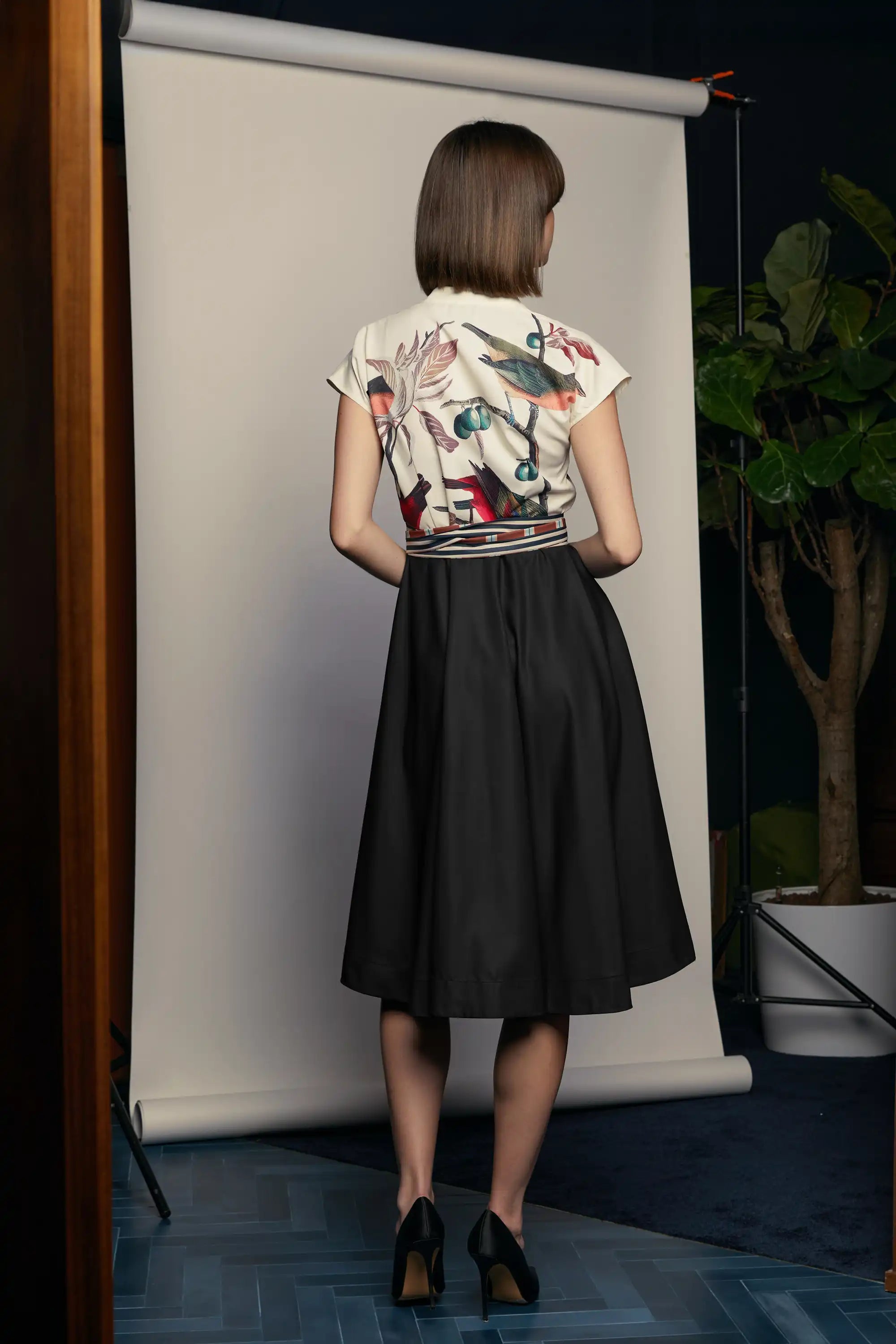 Back view of the HAVEN Surplice Fit & Flare Dress in the Painted Finch print. The cream bodice showcases a vivid bird and foliage illustration that continues seamlessly across the back, paired with a black flared skirt. The waist is accented with a striped sash belt, adding structure to the softly flowing silhouette.