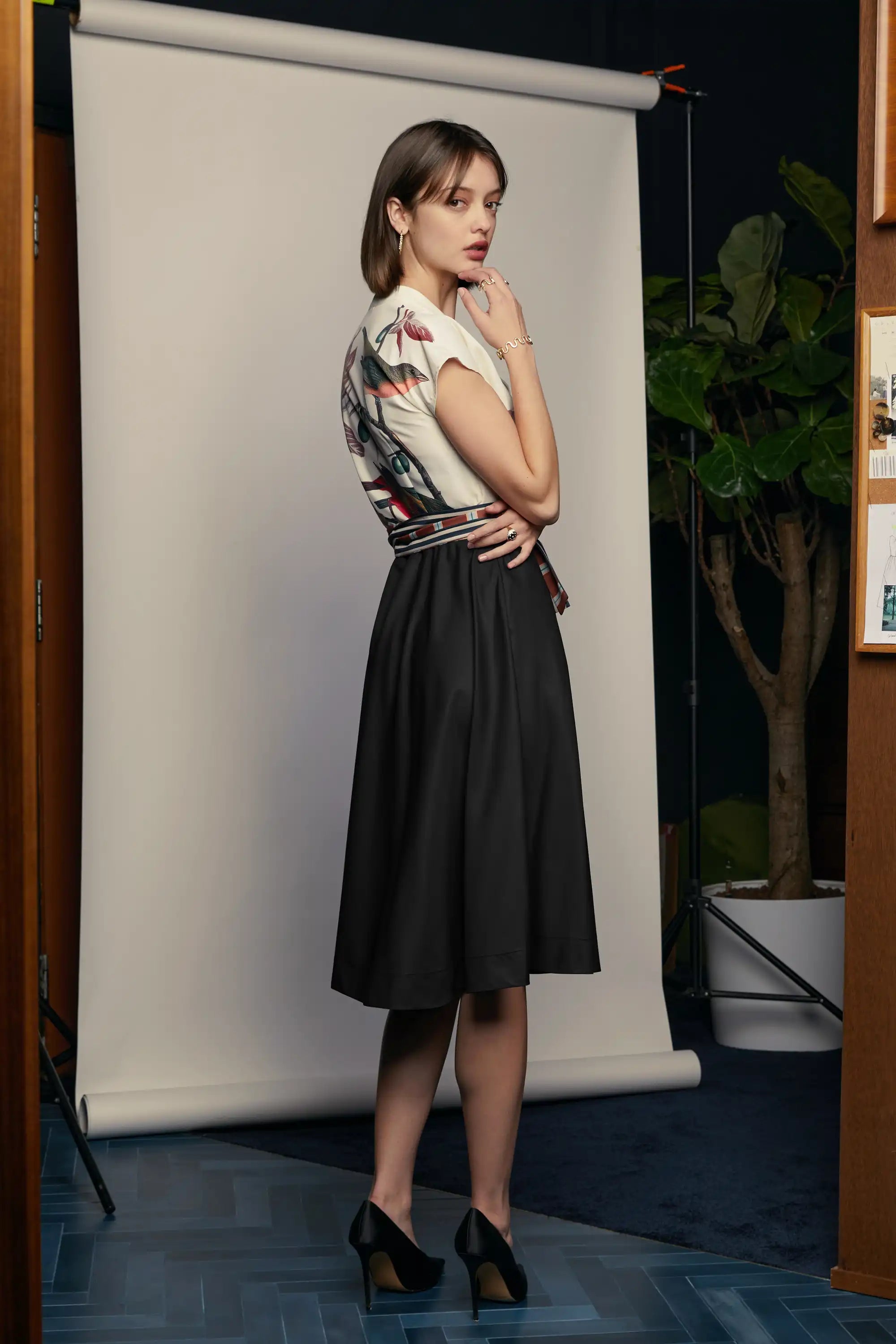 Close-up of the HAVEN Surplice Fit & Flare Dress in the Painted Finch print. The cream surplice bodice features a detailed bird and branch illustration with gold buttons along the neckline. A striped sash belt in shades of brick, navy, and teal is tied at the waist, complementing the black flared skirt and adding definition to the silhouette.