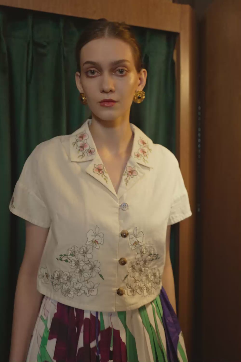 Close-up video of a model wearing a cropped Cuban shirt embroidered with botanical motifs, styled with pleated iris-print twirl skorts. Captured in warm studio lighting with a velvet curtain backdrop.