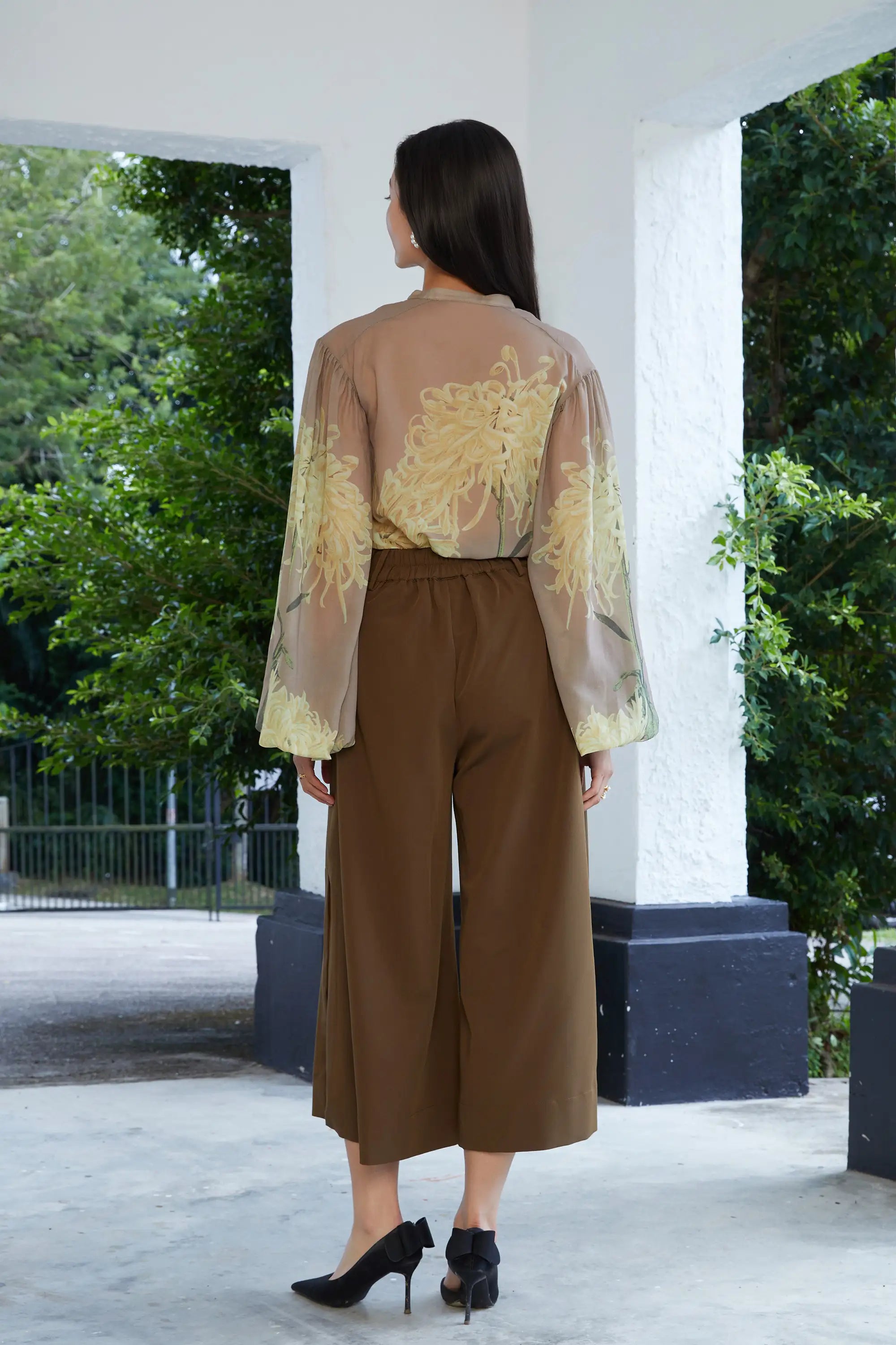 Woman wearing ABIDE sheer floral blouse with brown wide-leg slit pants outdoors (back view)
