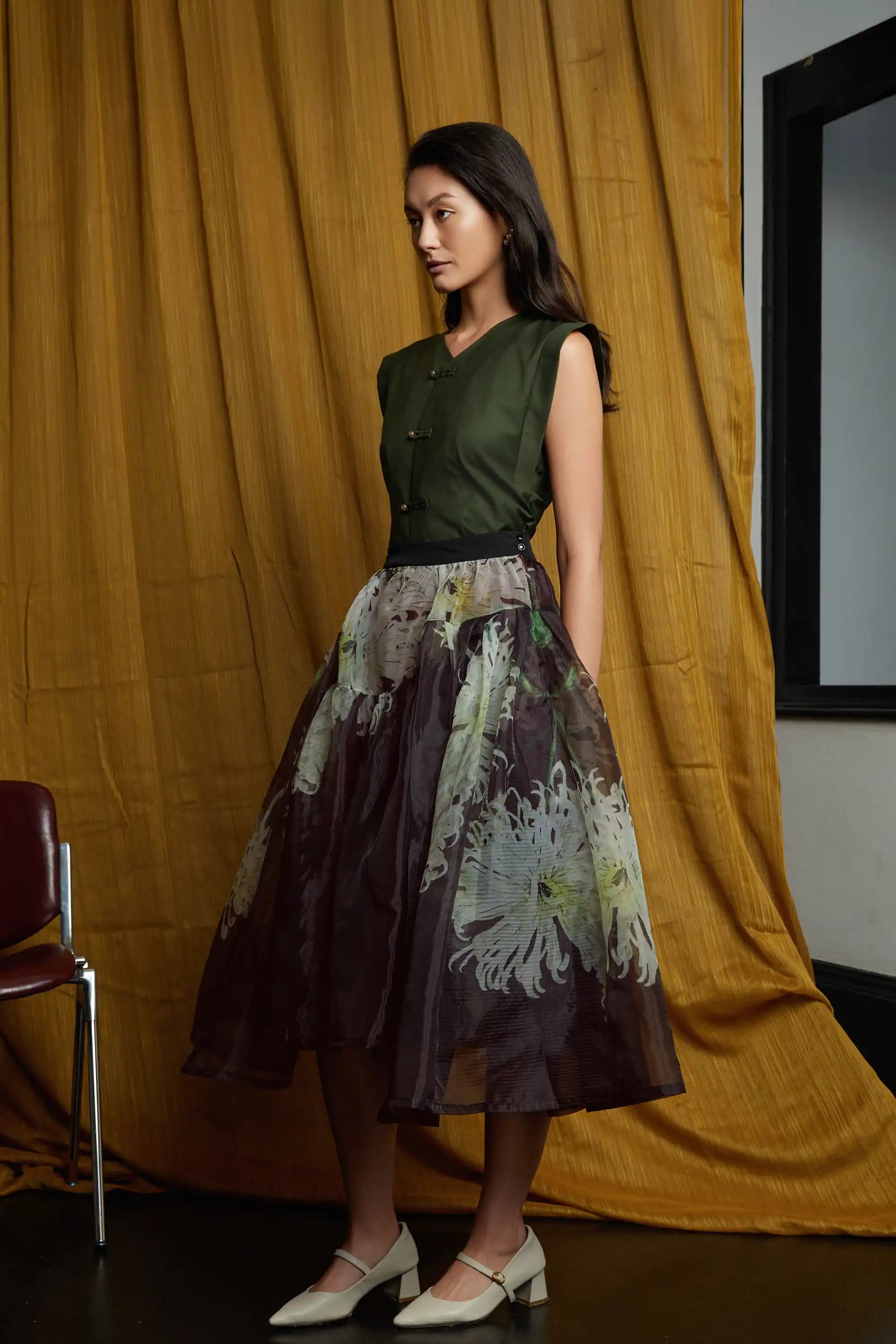 Deep green structured top with clean lines and subtle pankou closures, paired with a sheer floral overlay skirt, highlighting a cinched waist, soft volume, and fluid, airy movement