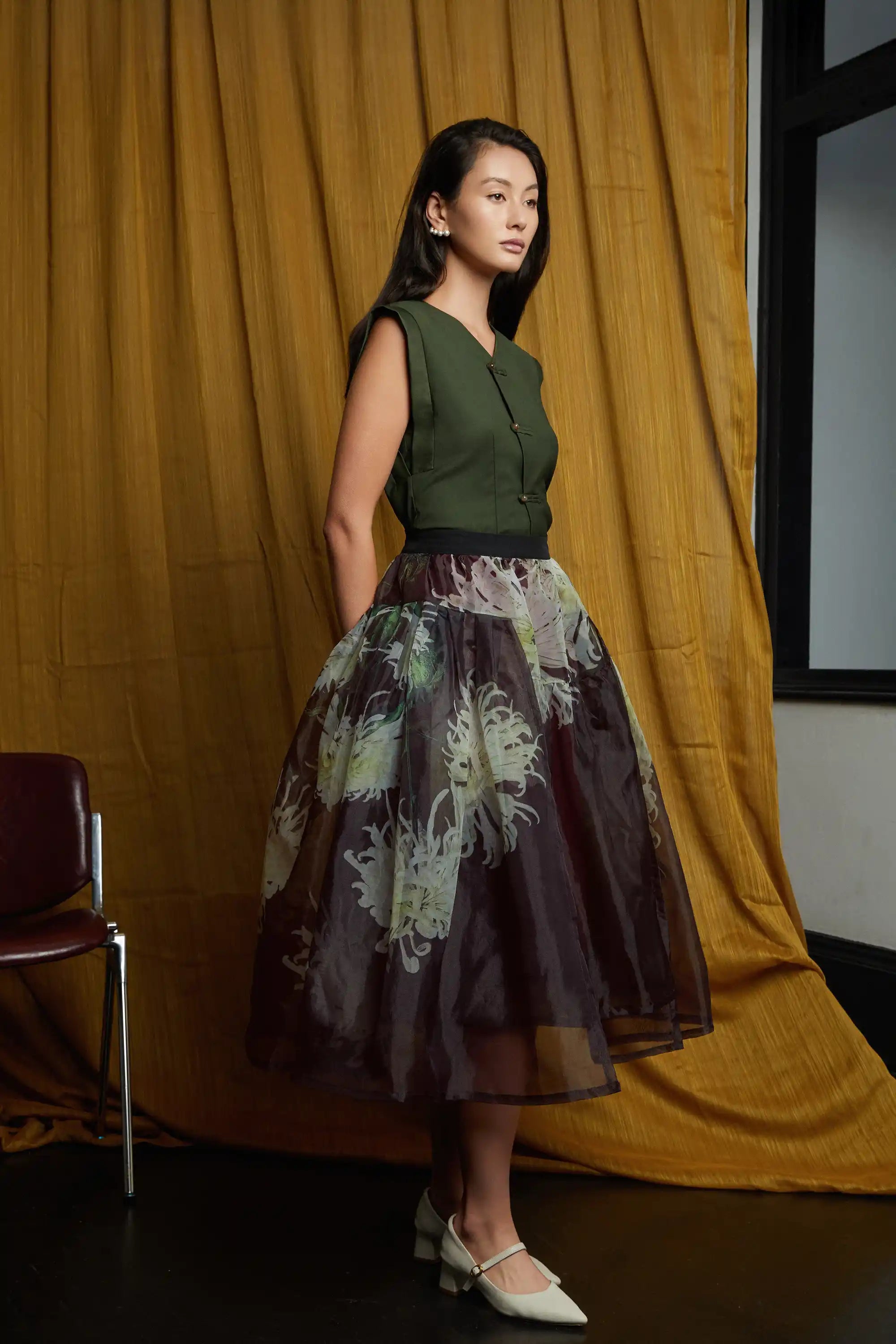 Deep green structured top with clean lines and subtle pankou closures, paired with a sheer floral overlay skirt, highlighting a cinched waist, soft volume, and fluid, airy movement