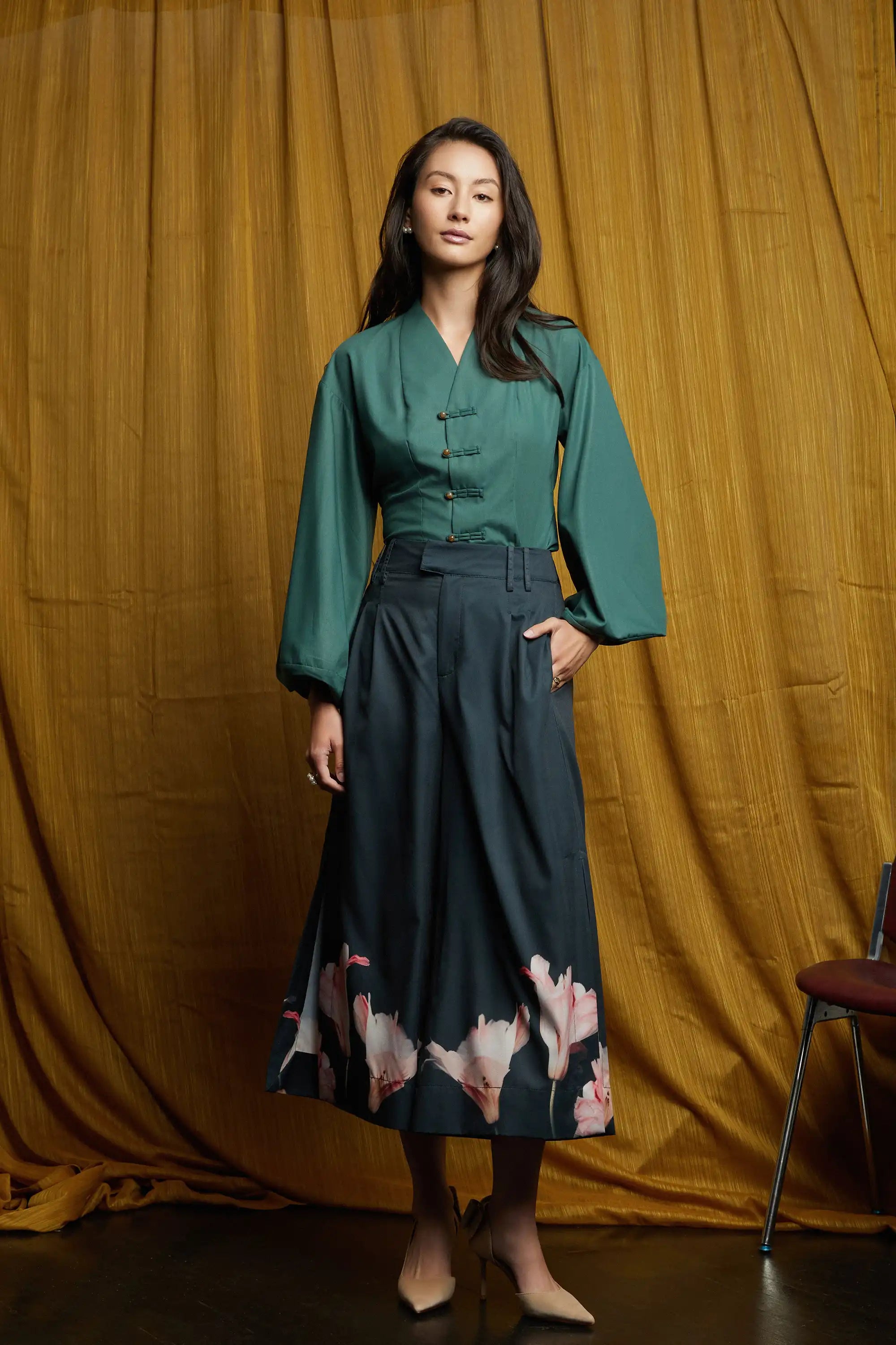 Full-length look featuring deep green long-sleeve top with pankou button closures and softly voluminous sleeves, paired with dark pleated slit pants with wide-leg silhouette and pink floral placement print at the hem