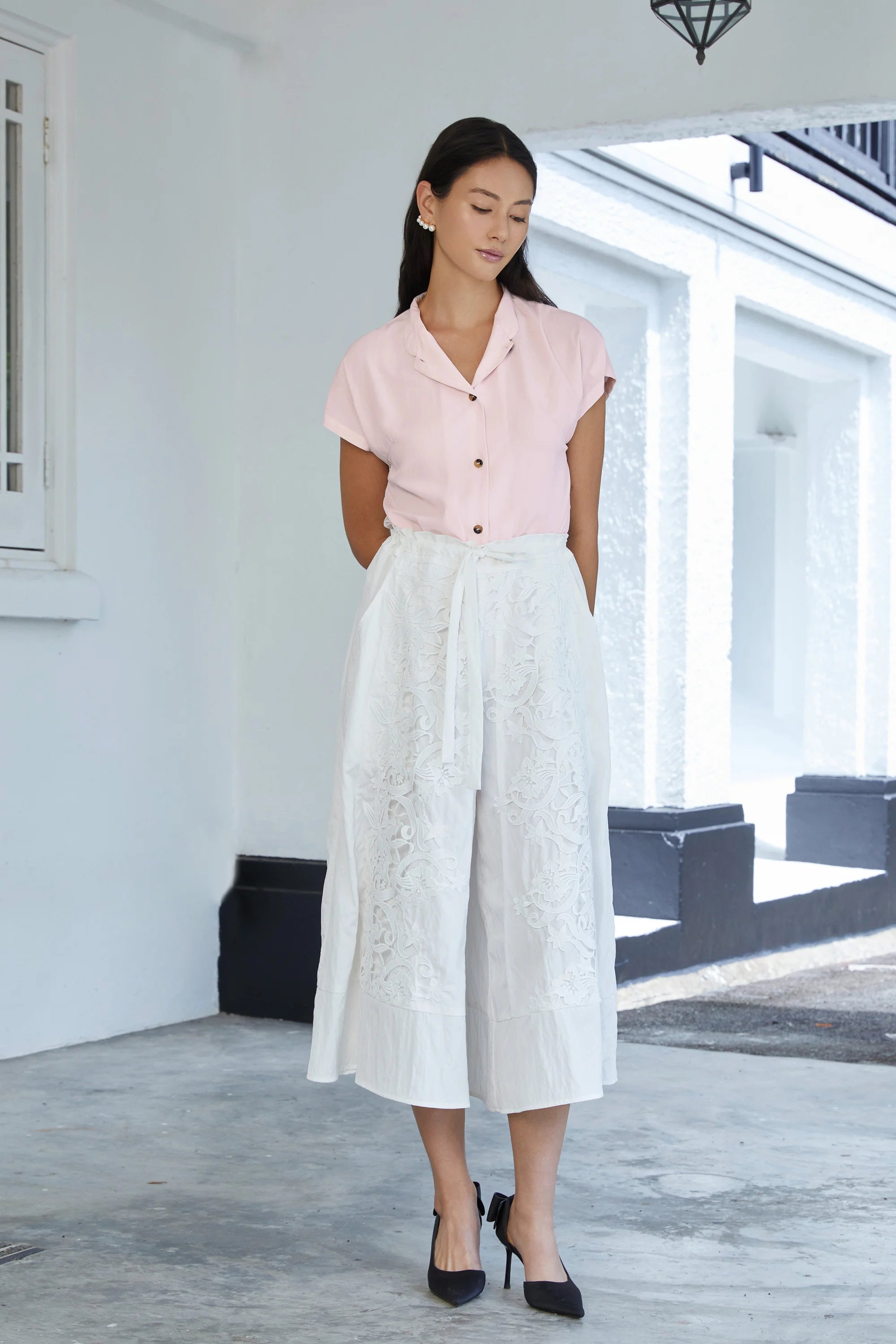Woman wearing blush pink short-sleeve blouse with button front, paired with white lace wide-leg palazzo pants with drawstring waist