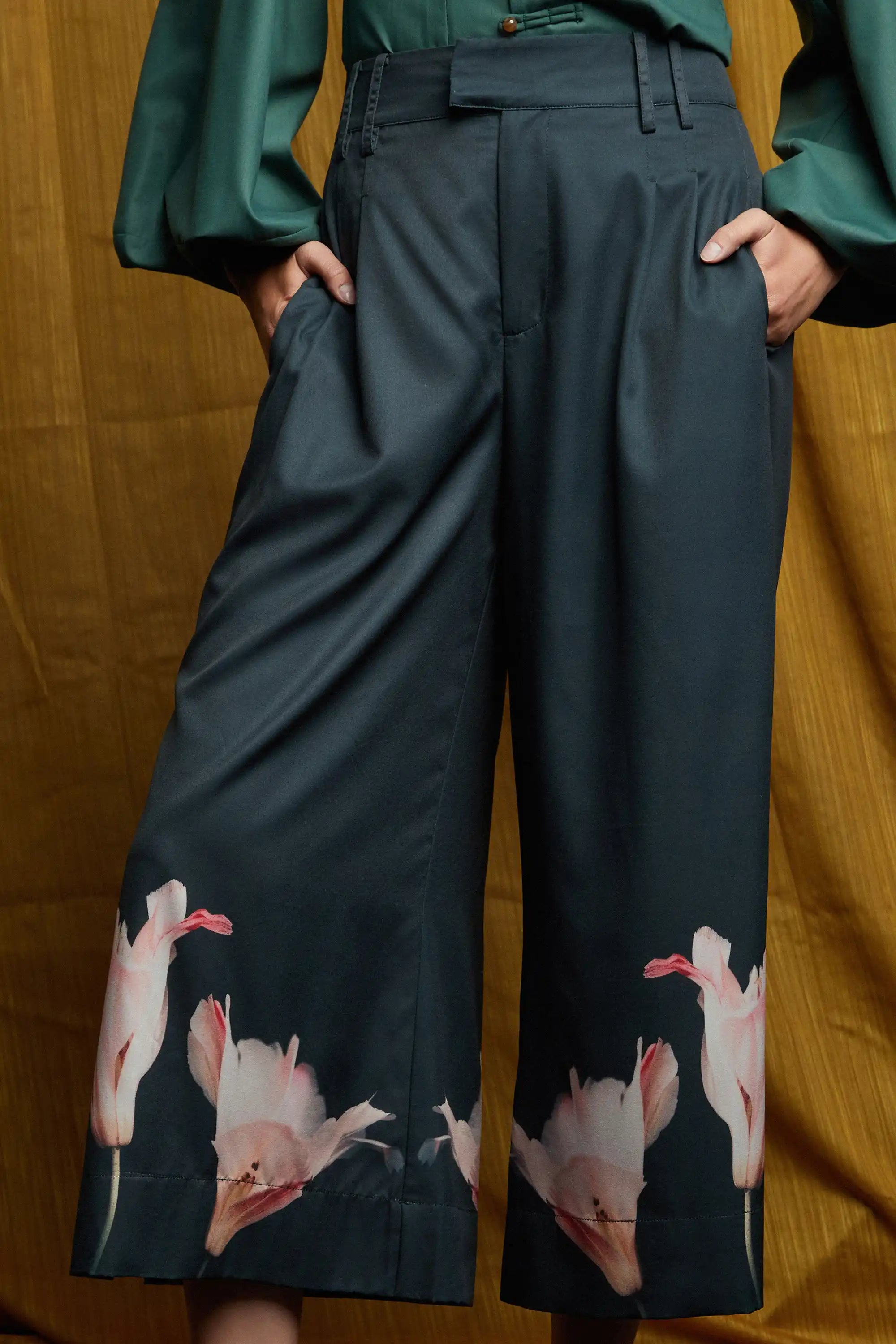Close-up of dark pleated slit pants highlighting high-waist structure, soft front pleats, and side pockets, finished with pink floral placement print along the hem and a fluid, wide-leg silhouette