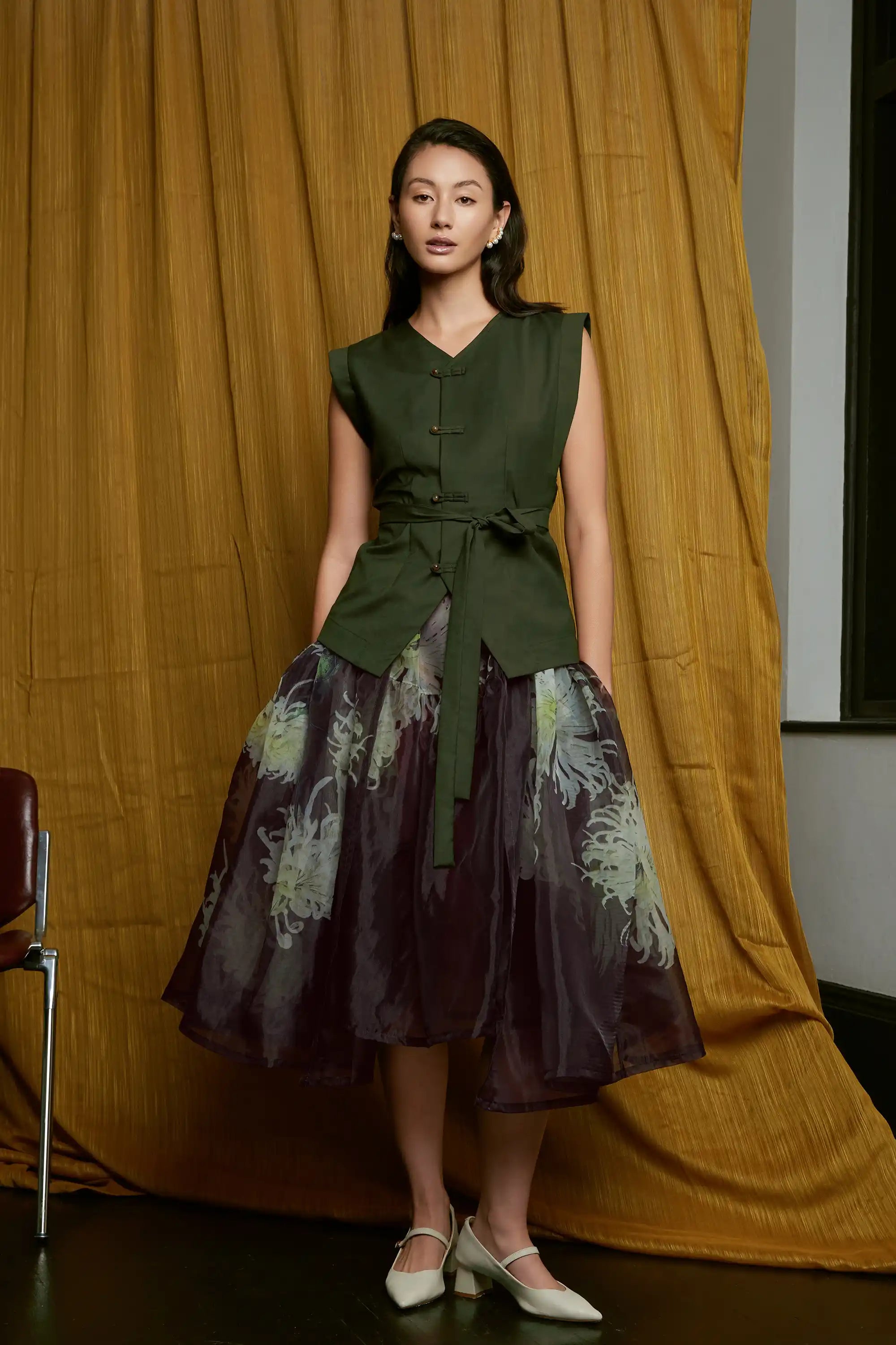 Full front view of model wearing deep green structured top with pankou button closures and waist sash tie, paired with a sheer floral overlay skirt, showcasing a cinched silhouette, layered transparency, and soft flowing movement