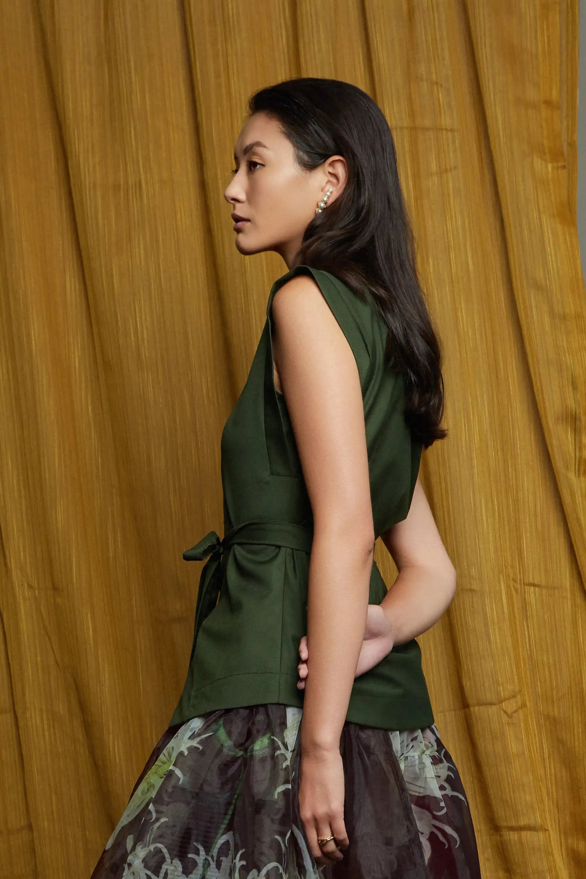 Deep green structured top with clean lines and subtle shoulder structure, styled with a sheer floral skirt, showcasing fluid drape and airy volume from the side