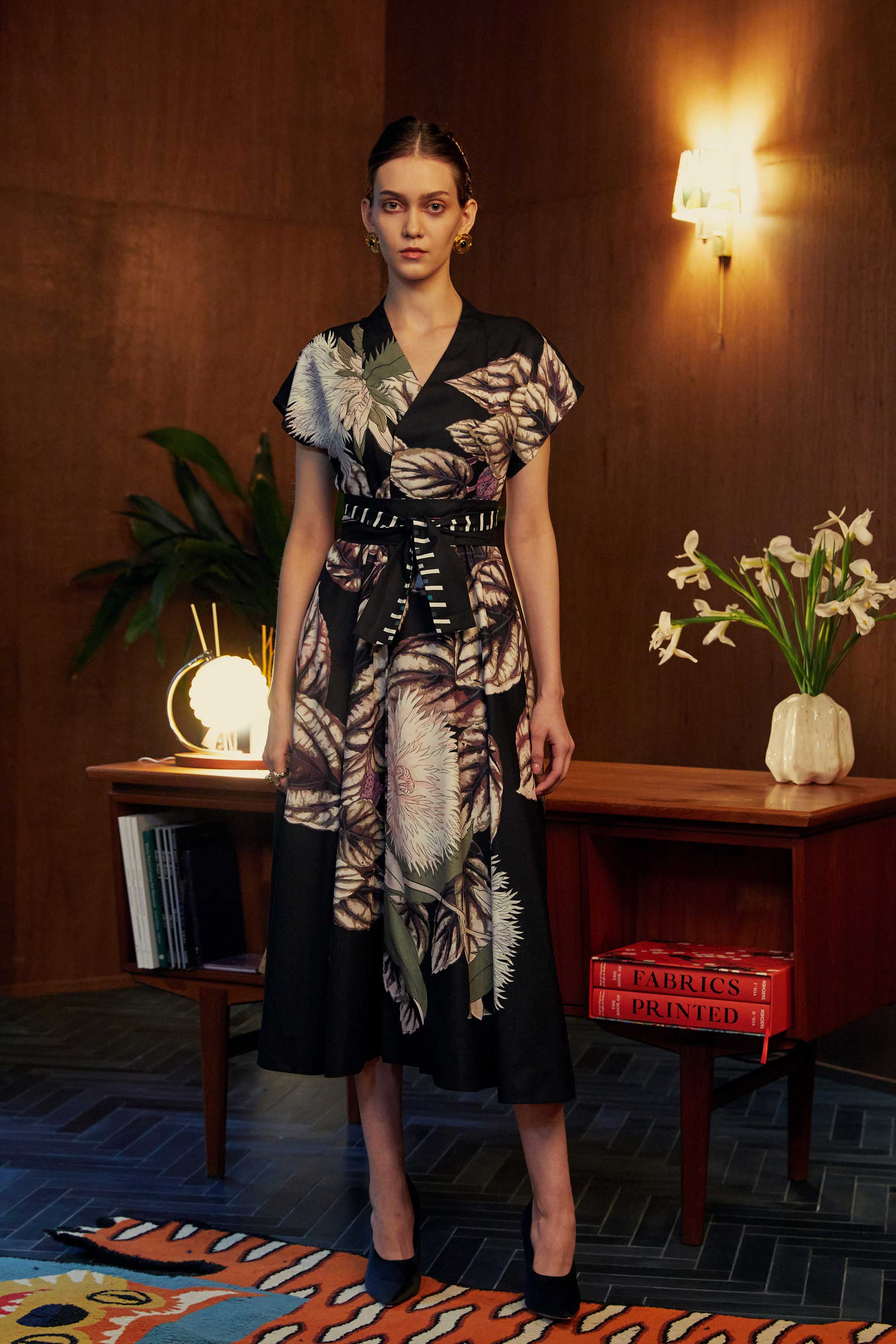 Model wearing the BEHOLD Blended Collar Wrap Midi Dress in Stokesia print, featuring layered leaf motifs in muted tones, styled with a cinched belt. Photographed in a wood-panelled room with warm ambient lighting and mid-century furniture.