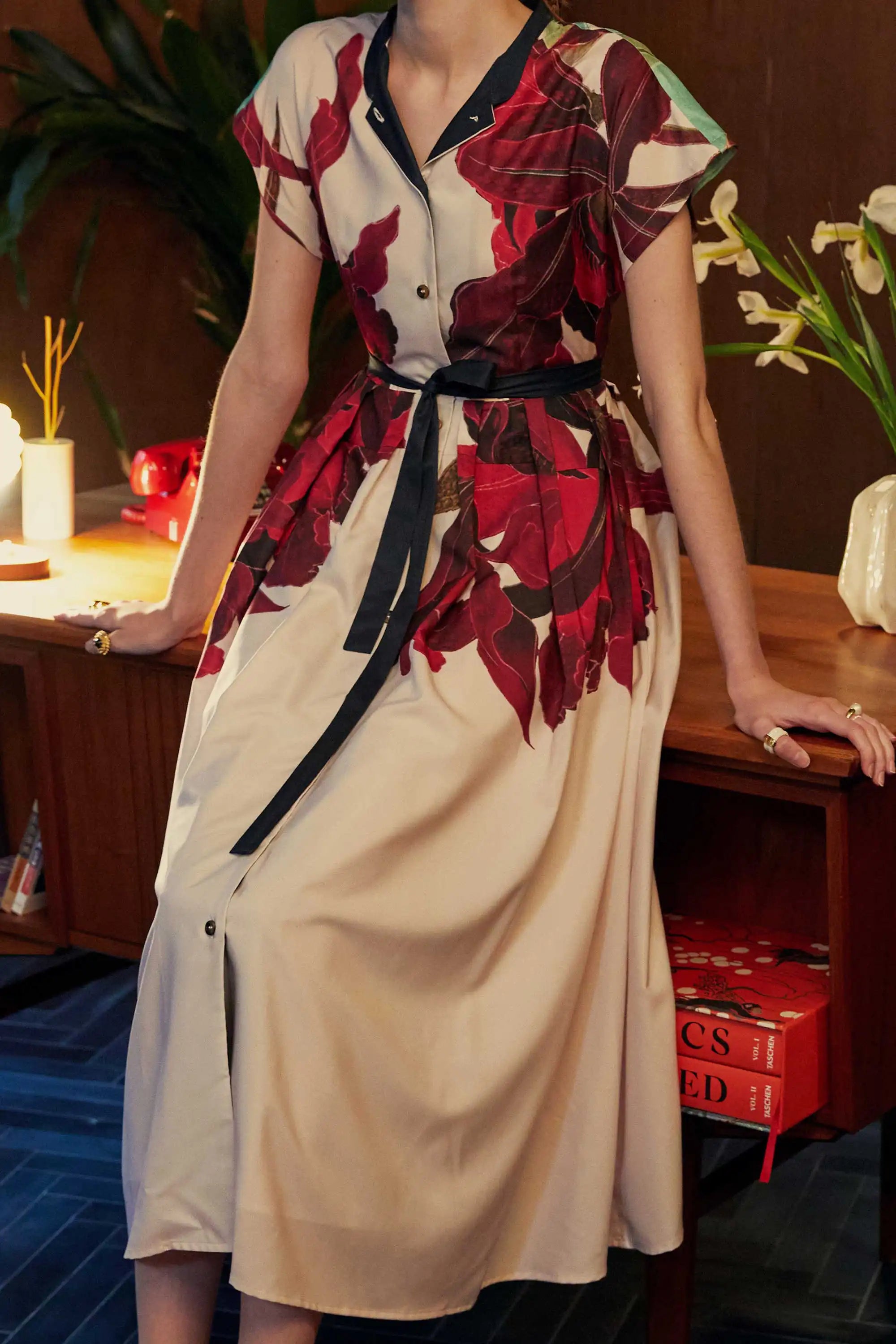 Close-up of the BEHOLD Chinese Collar Fit & Flare Maxi Dress in Laelia print, highlighting the oversized crimson orchid motifs against a cream base, with a black sash at the waist. Shot indoors with wood-panelled furniture, a tiger-striped rug, and ambient lighting