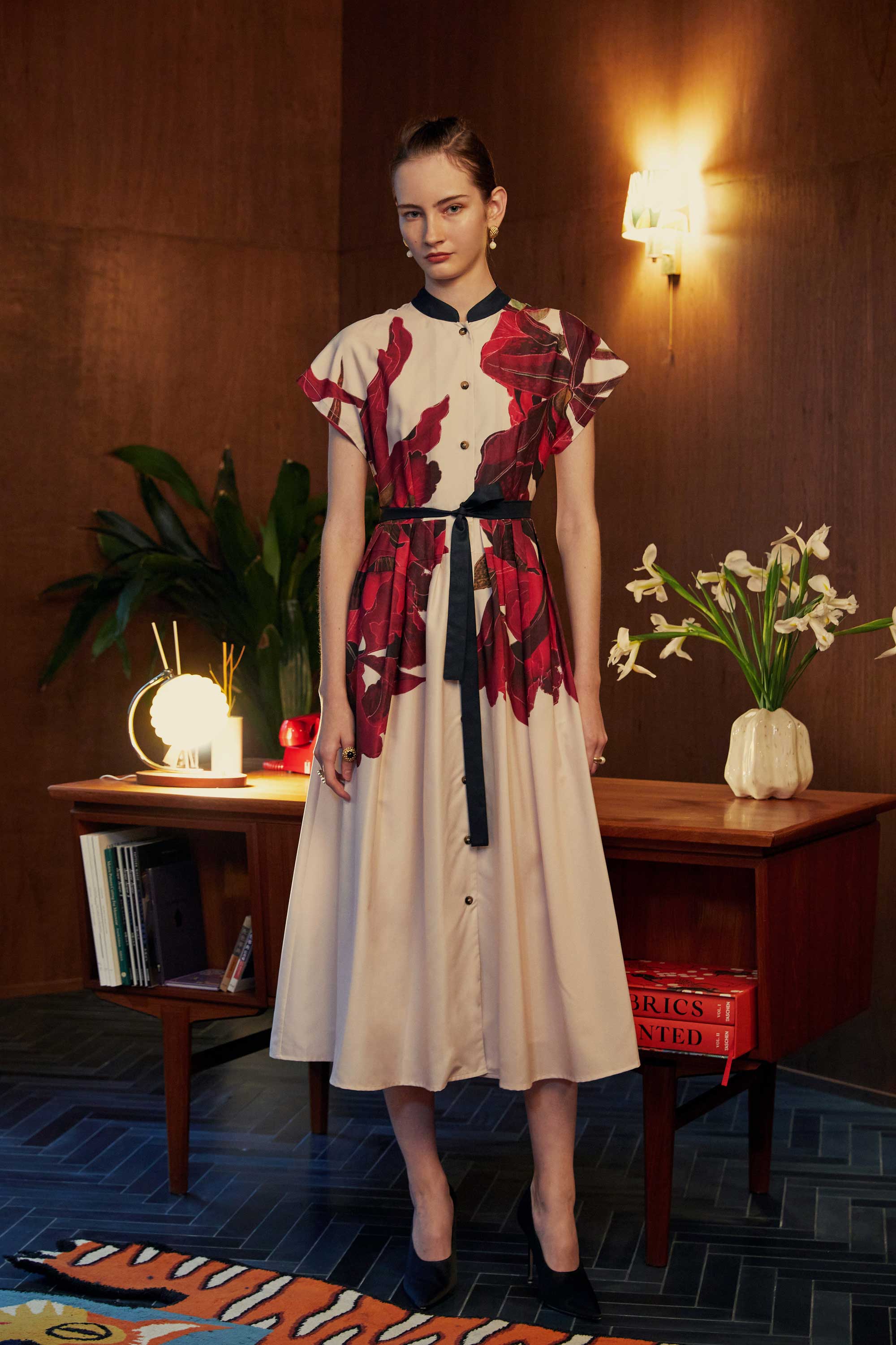 Model wearing the BEHOLD Chinese Collar Fit & Flare Maxi Dress in Laelia print, featuring bold orchid motifs in deep crimson and aubergine. Styled in a warm, wood-panelled room with ambient lighting, books, and floral decor.