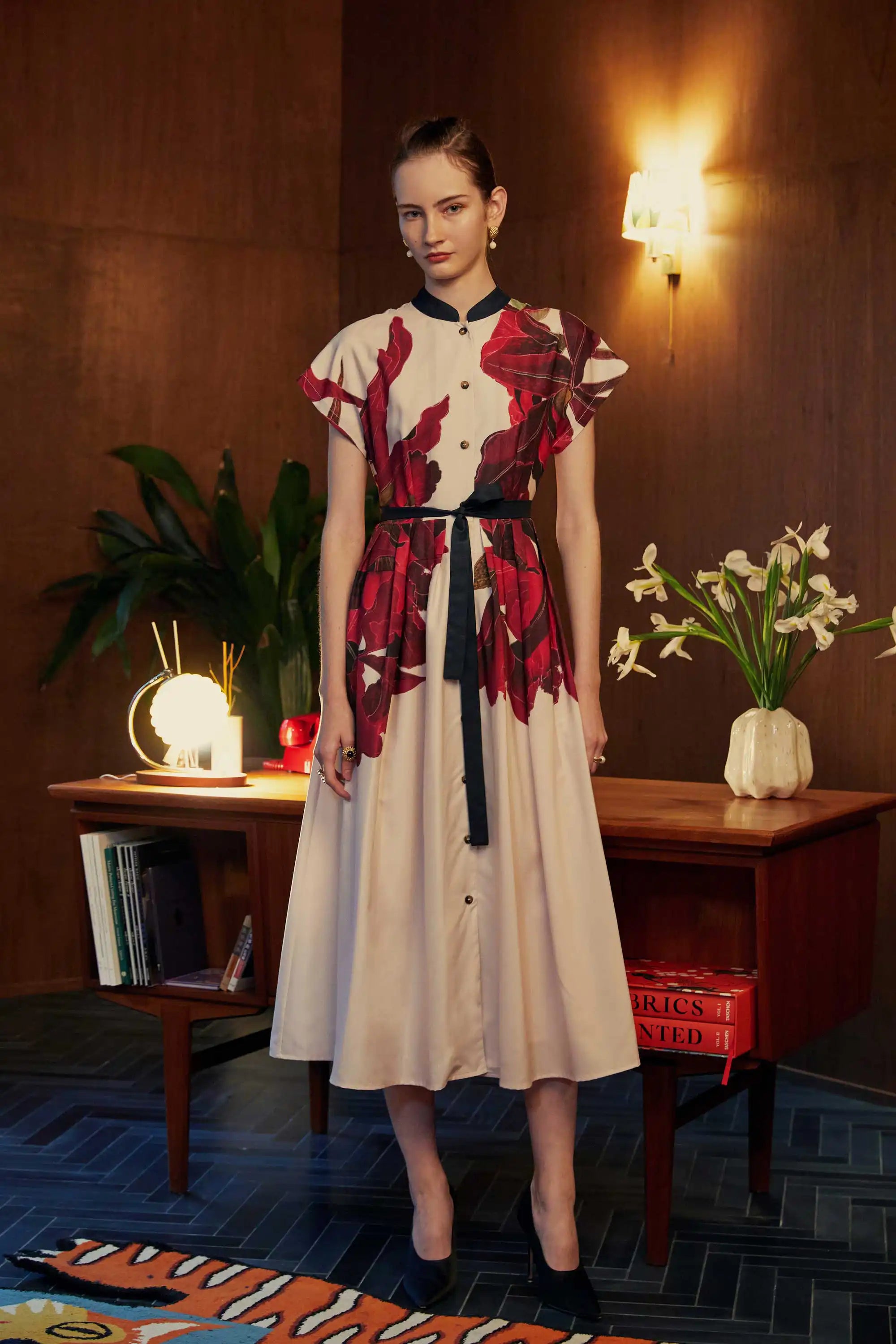 Model wearing the BEHOLD Chinese Collar Fit & Flare Maxi Dress in Laelia print, featuring bold orchid motifs in deep crimson and aubergine. Styled in a warm, wood-panelled room with ambient lighting, books, and floral decor.