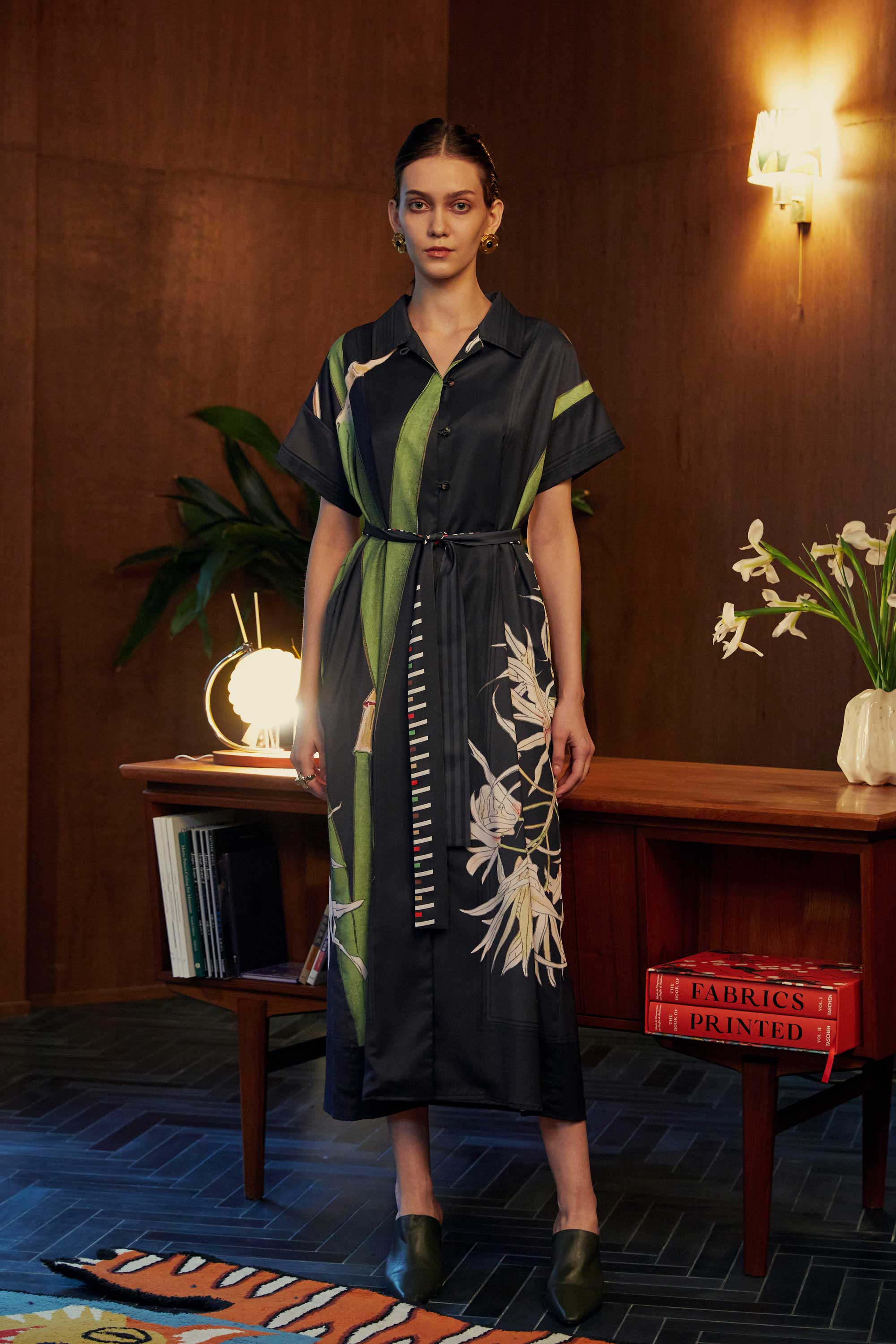 Model wearing the BEHOLD Cuban Maxi Dress in Brassavola Cucullata orchid print, black and green maxi shirt-dress with belted waist and floral motifs, styled in a wood-paneled indoor setting.