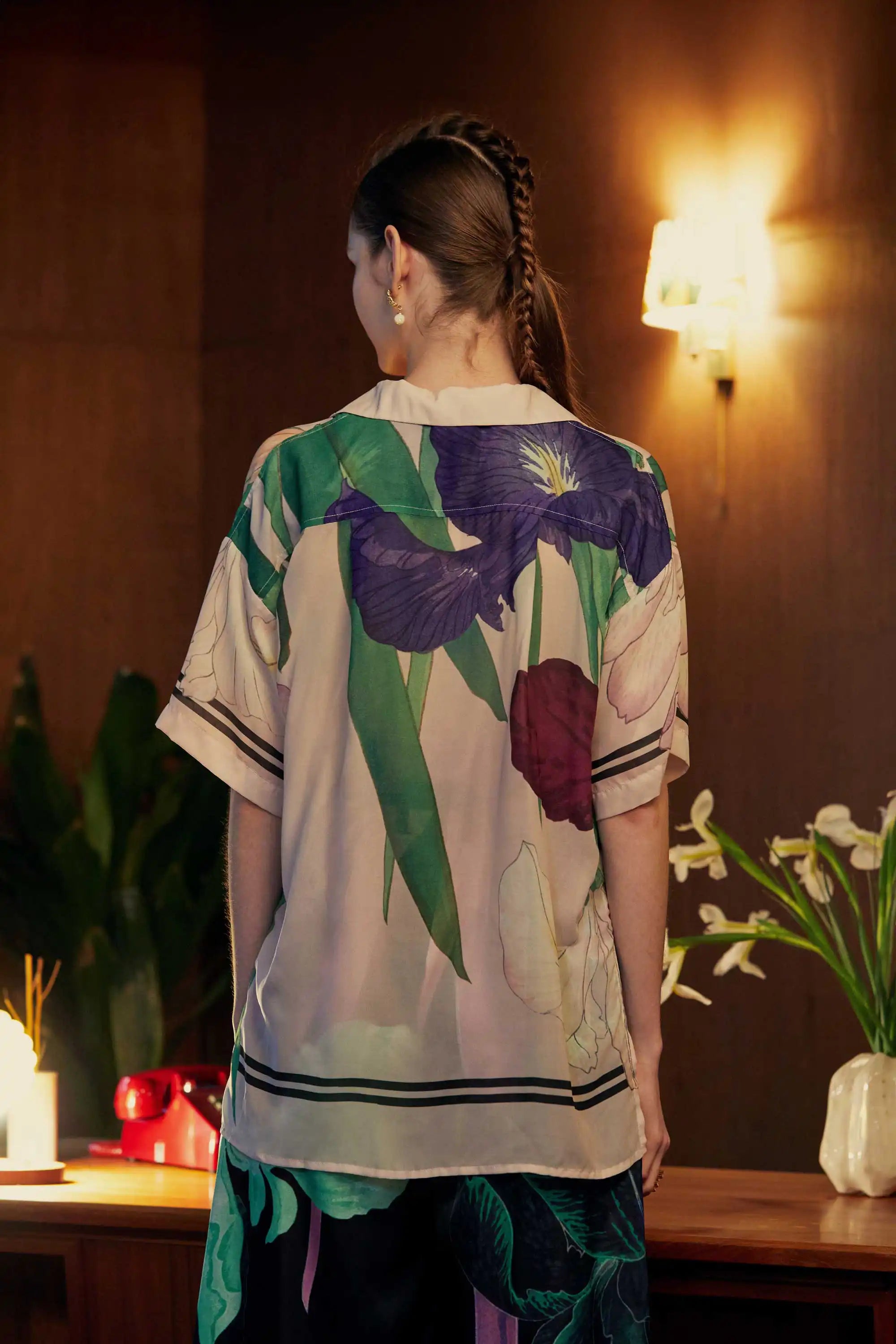 Back view of an oversized iris print Cuban shirt, showcasing large painterly florals in plum and violet against a blush-toned base, photographed in a mid-century modern interior with soft lighting and leafy plants.