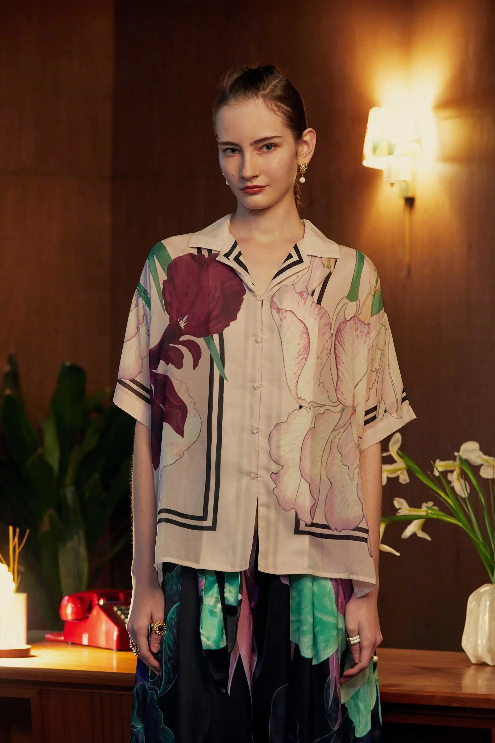 Model wearing an oversized floral Cuban shirt with bold iris motifs in pink and plum, styled in a mid-century interior with warm wood tones, tropical plants, and soft ambient lighting.