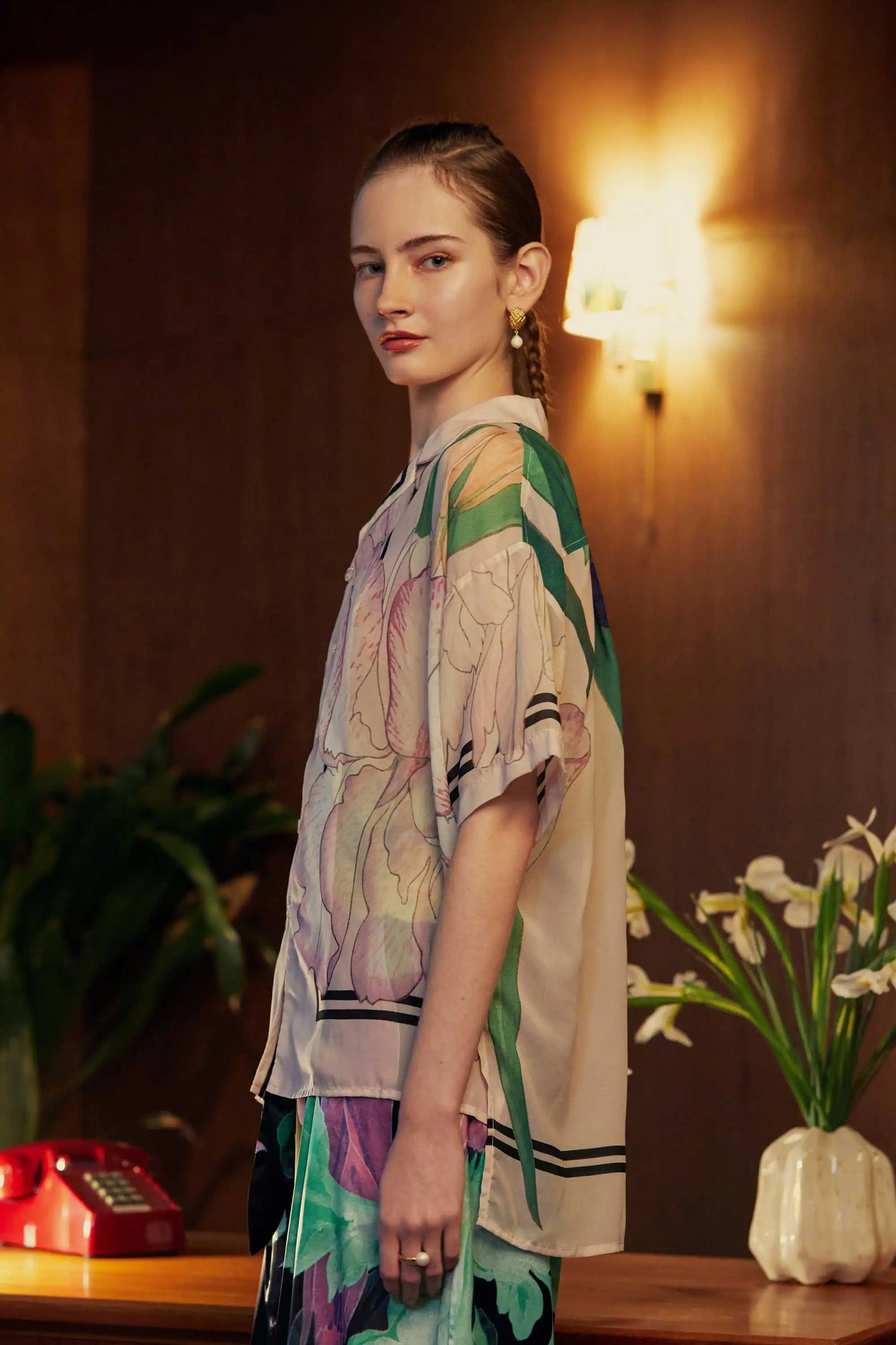 Model in an oversized Cuban shirt with vivid iris florals, shown from the left side to highlight the sleeve drape and botanical print, set against warm wood paneling and mid-century accents.