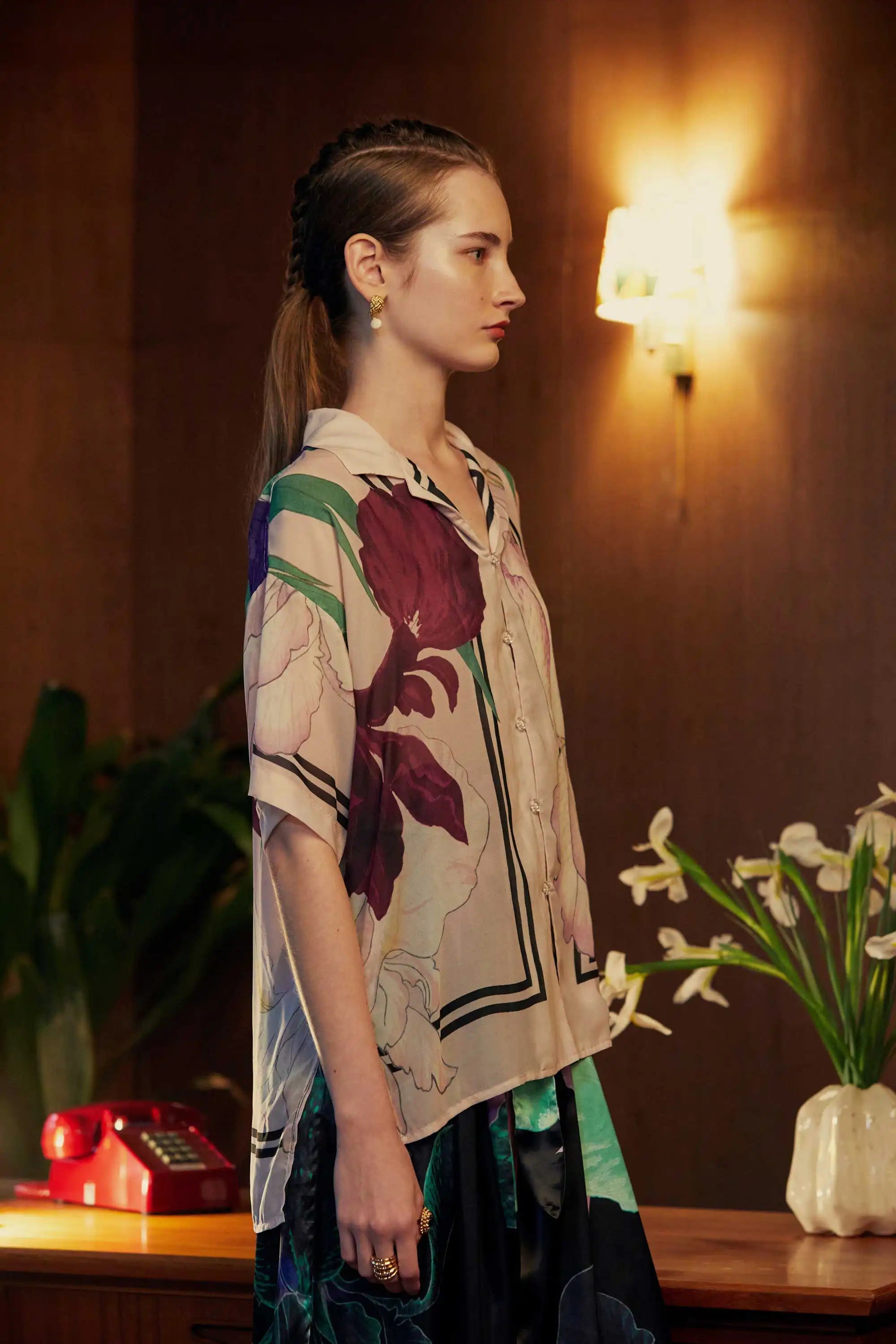 Right side view of a floral Cuban shirt in soft blush tones with oversized iris motifs in plum and cream, styled in a warmly lit room with a retro lamp and vase of white lilies.