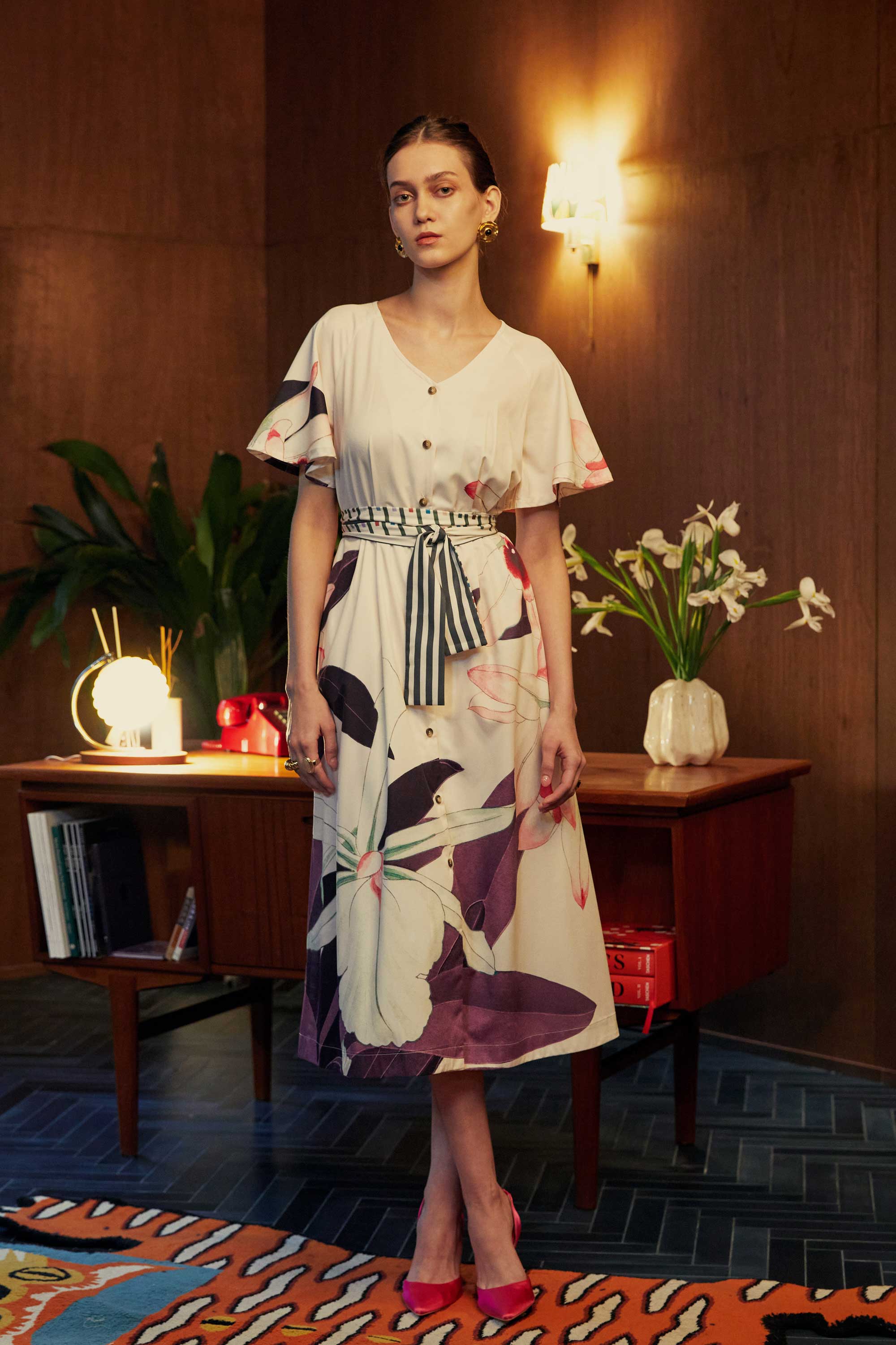 Model wearing the BEHOLD Flutter Sleeves Box Pleats Cinch Midi Dress in Dendrobium orchid print, featuring flutter sleeves, V-neckline, striped cinched waist, and cream base with bold orchid floral motifs, styled indoors with wood-paneled backdrop.