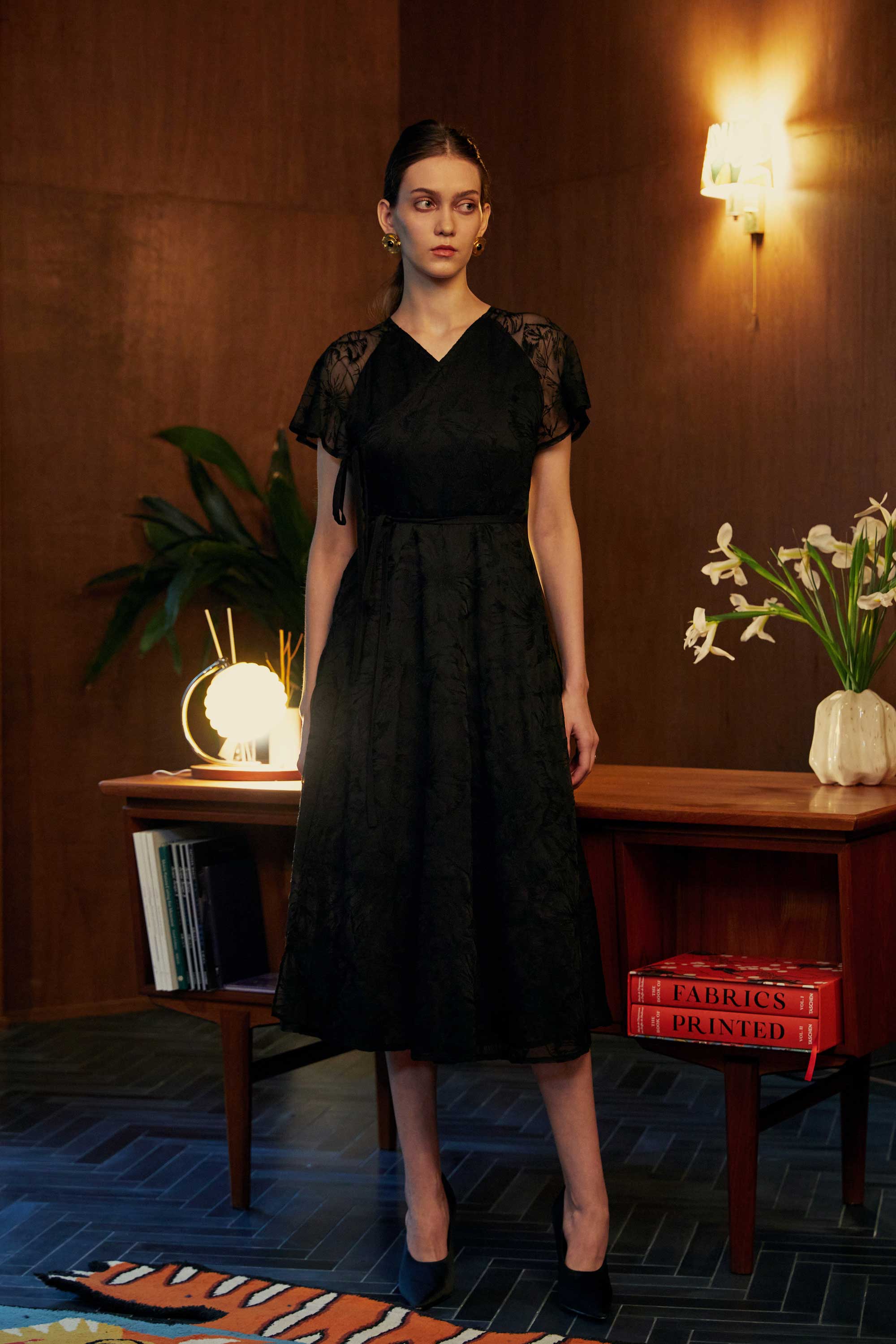 Model wearing the BEHOLD Flutter Sleeves Wrap Midi Dress in black lace, featuring V-neckline, flutter sleeves, and wrap silhouette, styled indoors with wood-paneled backdrop and warm lighting.
