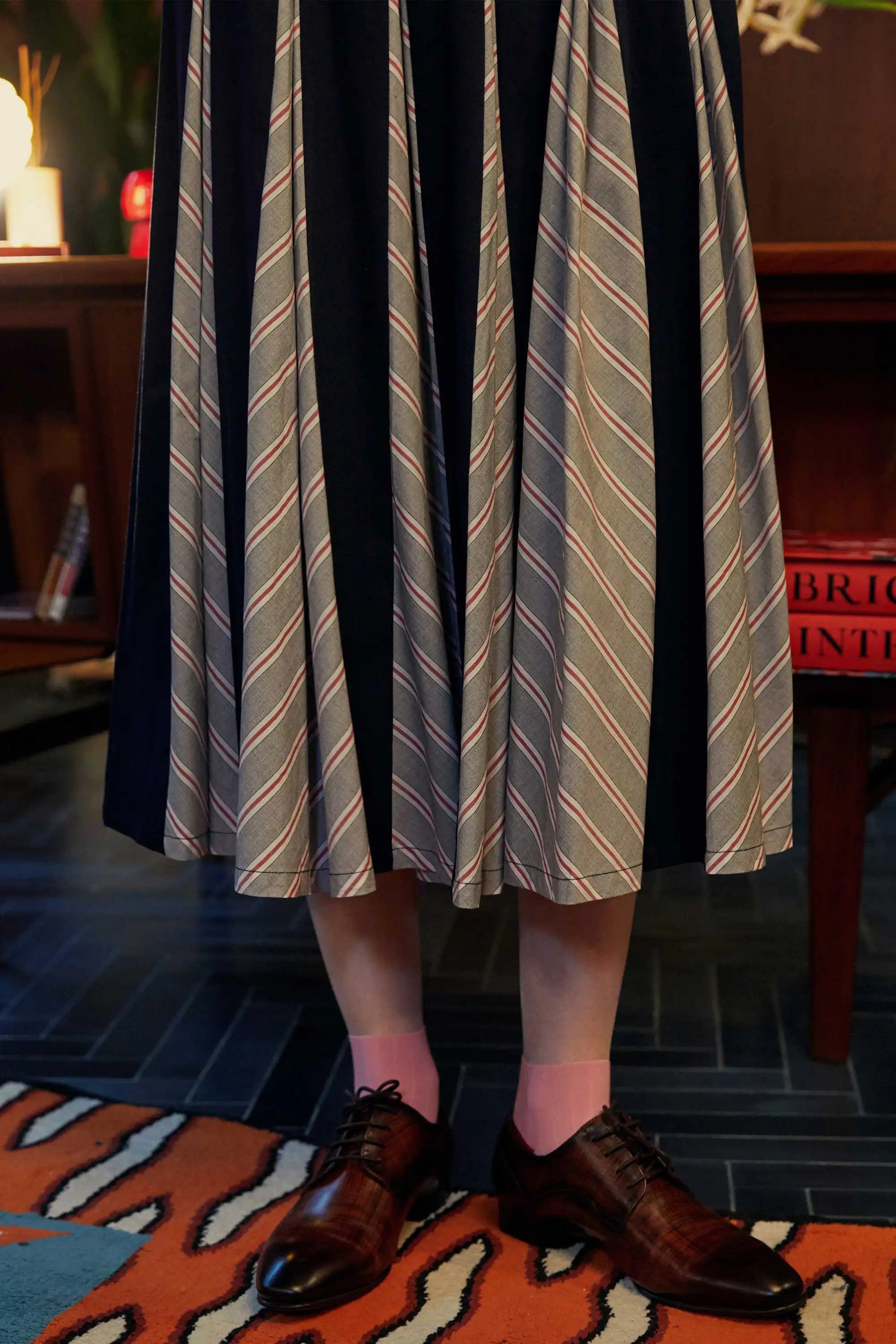 Close-up of the Godet Skirt in Navy Red, showcasing alternating chevron-striped godets and navy panels, styled with pink socks and oxford shoes on a tiger-striped rug.