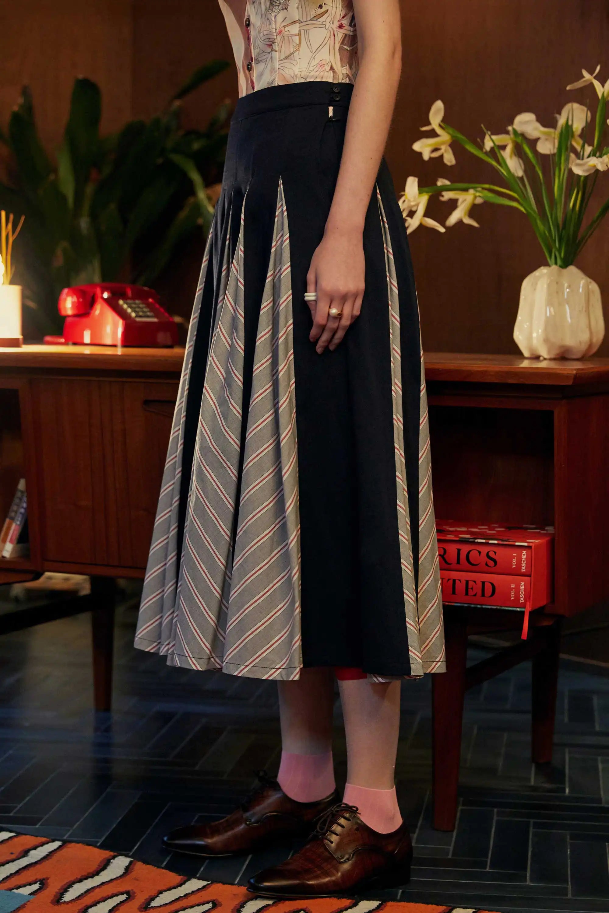 Side view of the Godet Skirt in Navy Red, featuring alternating navy panels and grey chevron-striped inserts, styled with pink socks and brown oxford shoes in a warm-toned room.