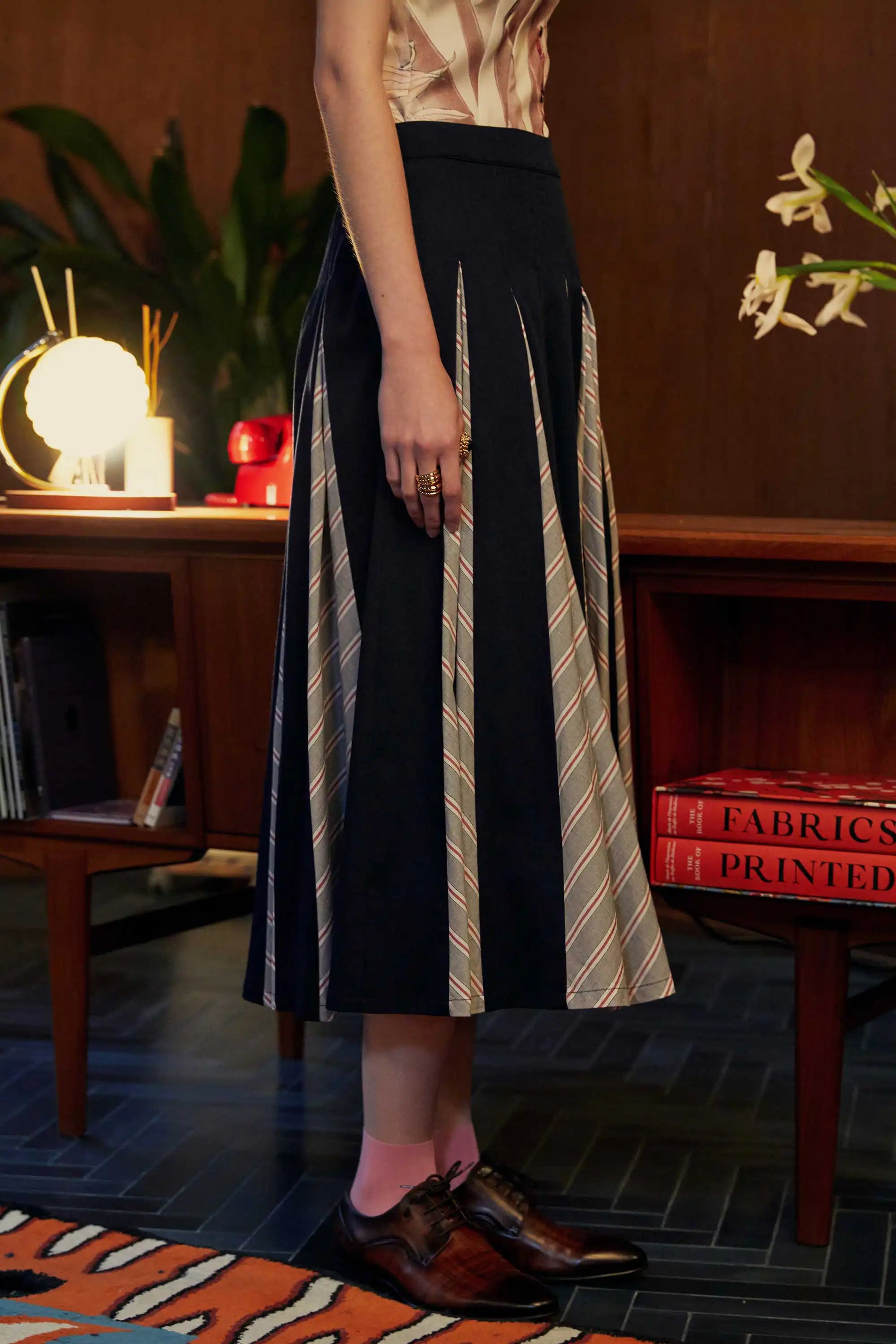 Alternate side view of the Godet Skirt in Navy Red, highlighting contrast godet panels in chevron-striped grey against navy, styled with brown brogues and pink socks in a wood-paneled room with books and floral decor.