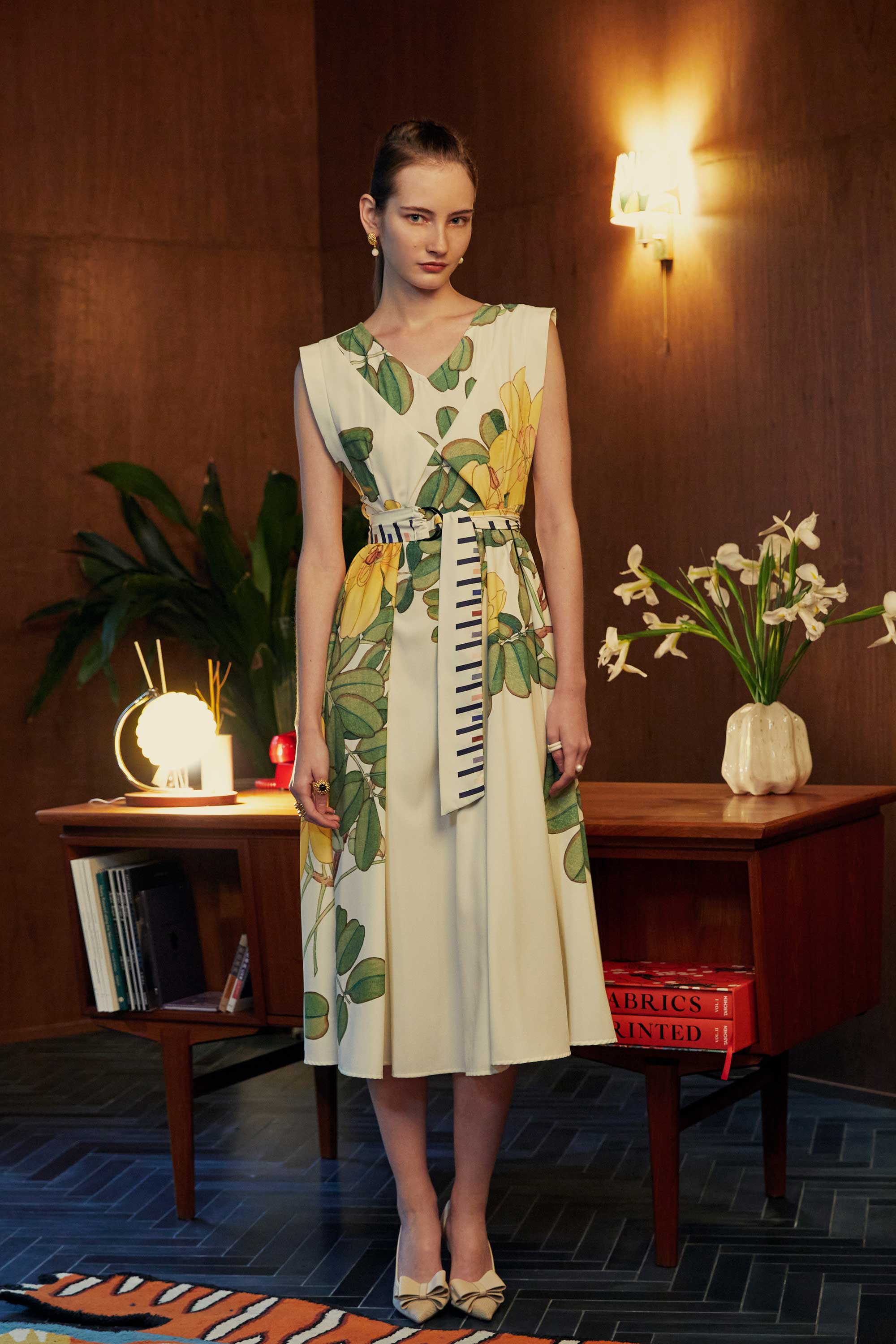 Model wearing the BEHOLD Origami Square Sleeve Dress with yellow allamanda floral print and striped obi belt, standing in a mid-century styled room with a wooden desk, flowers, and warm lighting.