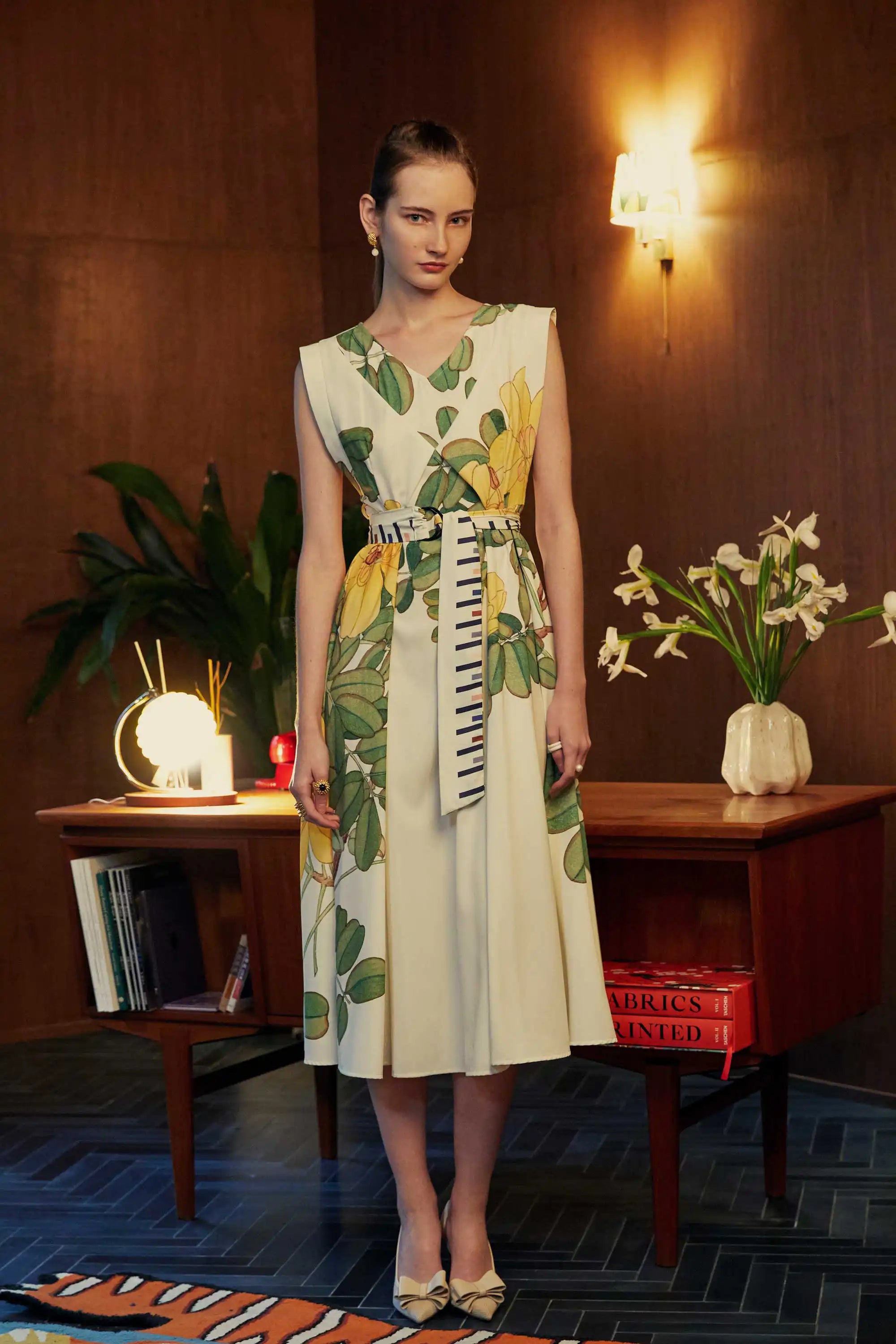 Model wearing the BEHOLD Origami Square Sleeve Dress with yellow allamanda floral print and striped obi belt, standing in a mid-century styled room with a wooden desk, flowers, and warm lighting.