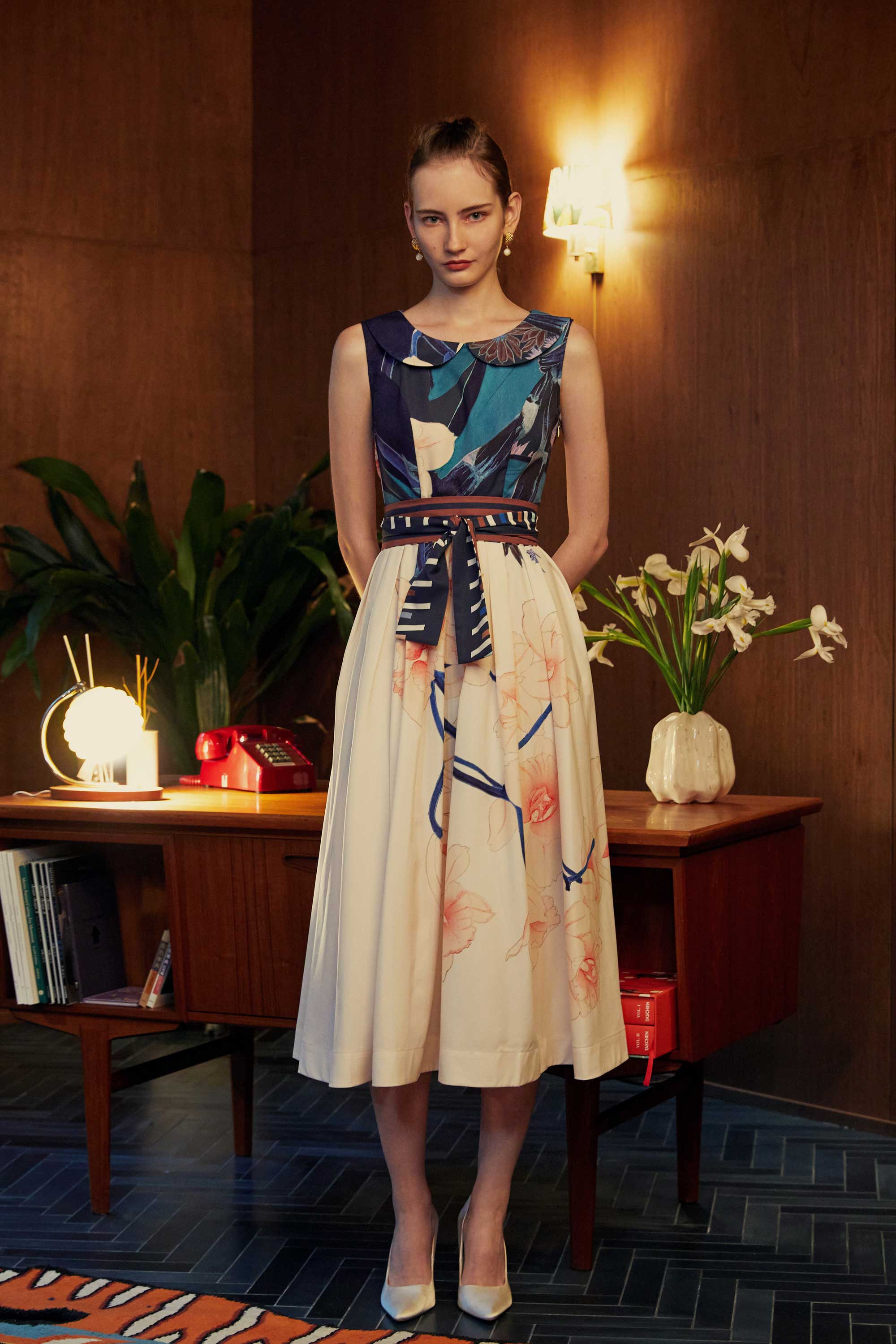 Model wearing the BEHOLD Peter Pan Fit-and-Flare Midi Dress in Dendrobium Chrysotoxum orchid print, featuring navy sleeveless bodice with Peter Pan collar, cream flared midi skirt with bold orchid floral motifs, and striped waistband, styled indoors with wood-paneled backdrop.