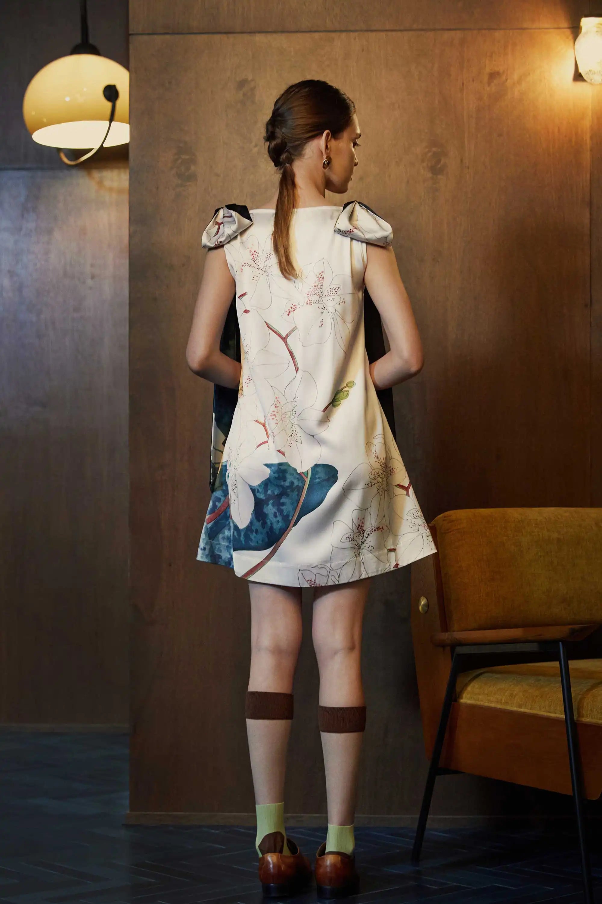 Back view of the BEHOLD Scarf Knee Dress in Moth Orchid print—cream A-line mini with scarf-tie shoulder straps and blue-green orchid motifs, styled indoors against wood-paneled walls.