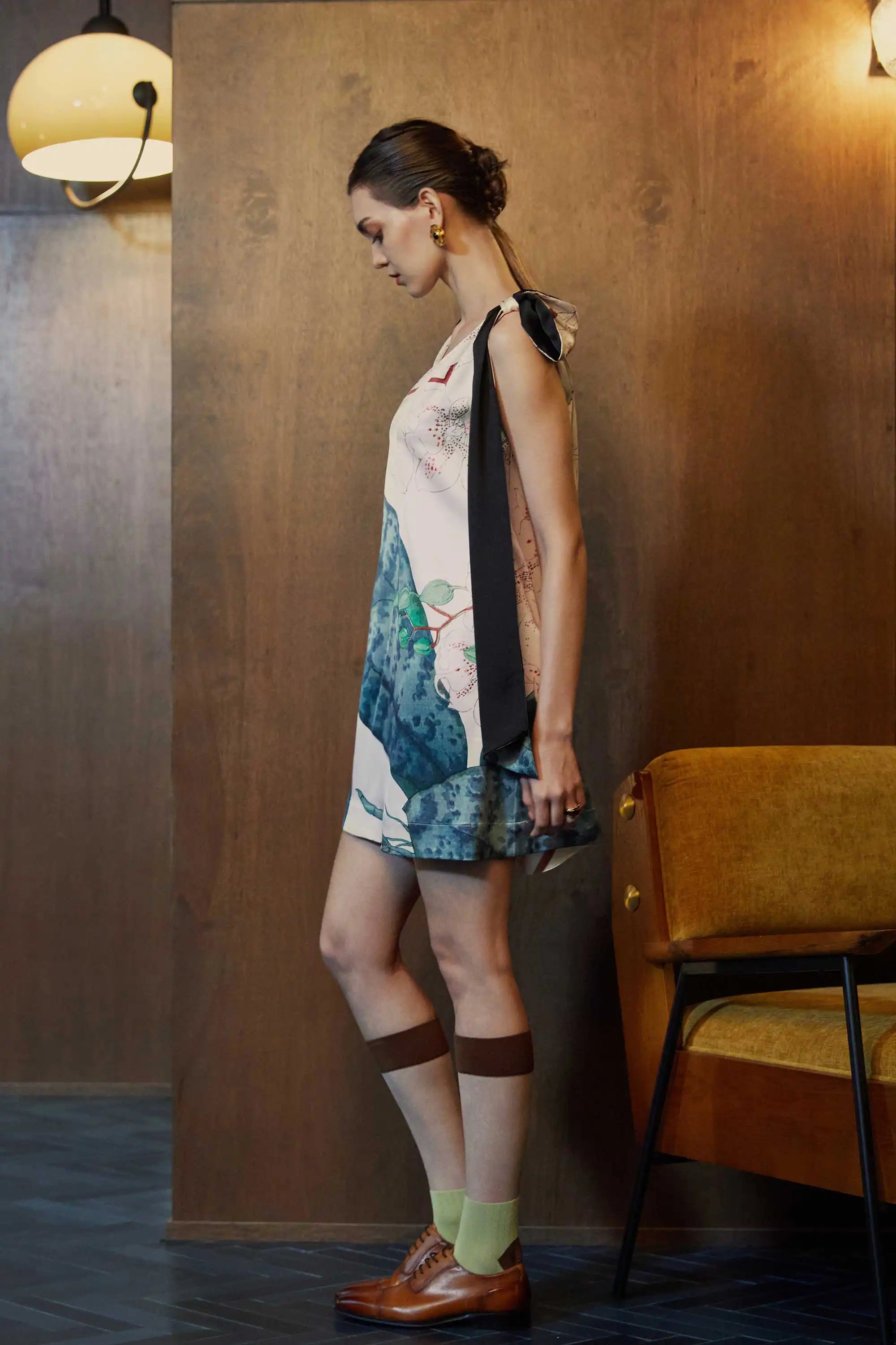 Side profile of model in the BEHOLD Scarf Knee Dress in Moth Orchid print—cream A-line mini with statement scarf-tie shoulder straps and blue-green orchid motifs, styled indoors against wood-paneled walls.
