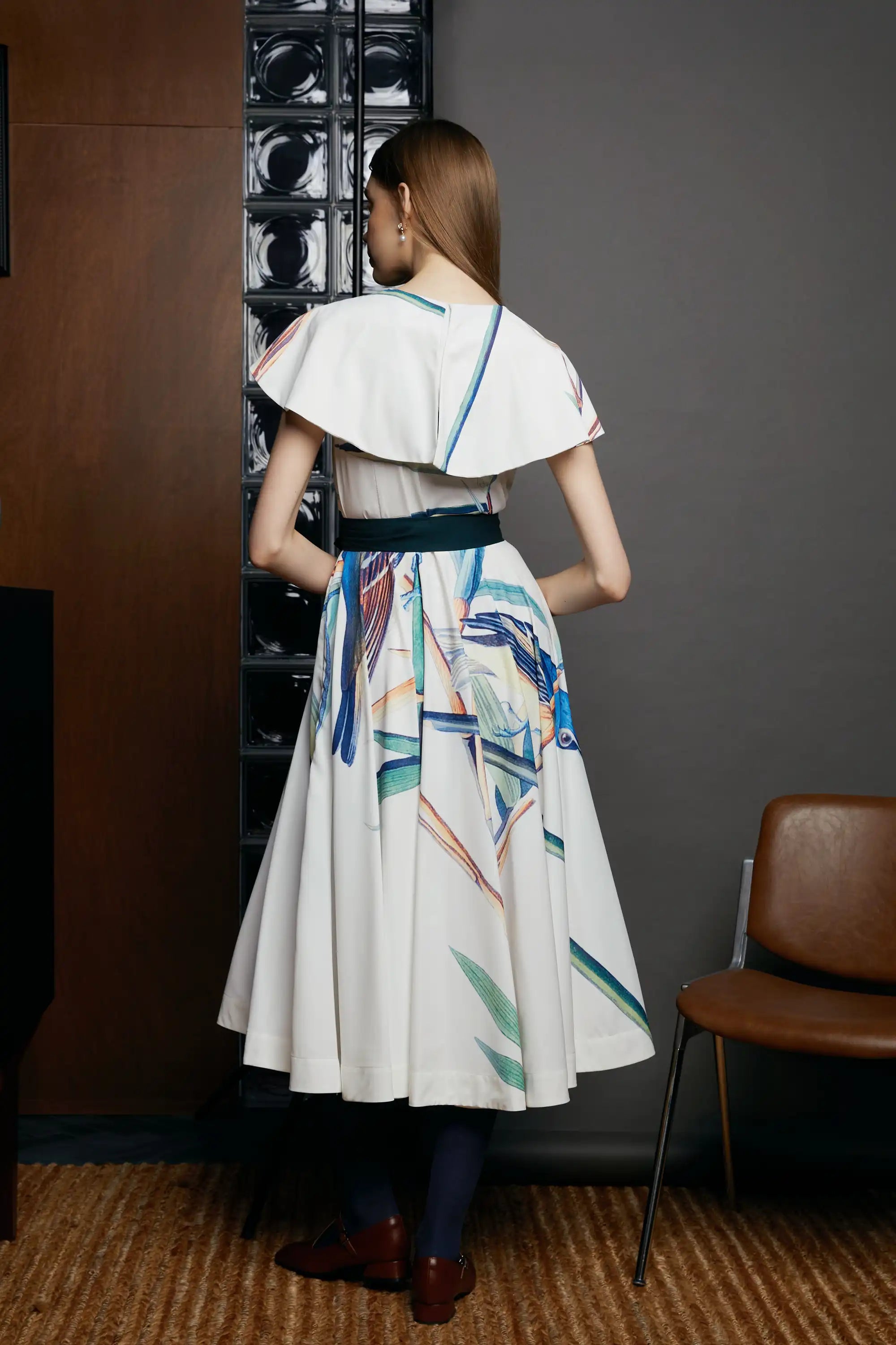 Back view of the HAVEN Blue-headed Vireo Caplet Collar Dress in cream with abstract blue brushstroke detailing. The dress features flutter sleeves, caplet collar, and a belted waist, styled in an indoor setting with a grey wall and brown chair.