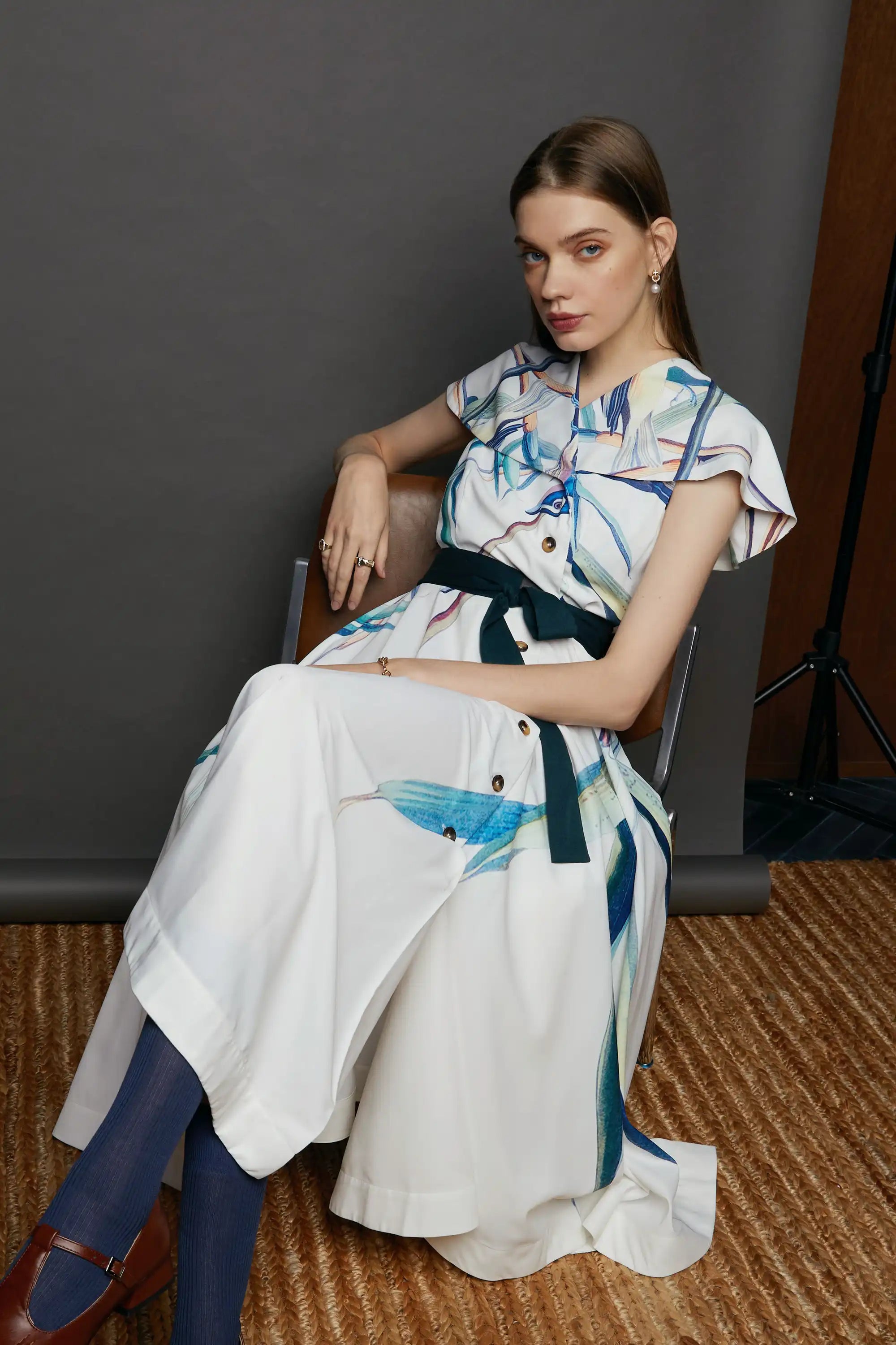 Model seated in a chair wearing the HAVEN Blue-headed Vireo Caplet Collar Dress in cream white with painterly teal and blue brushstroke bird motifs. The caplet collar, flutter sleeves, and dark waist tie are visible, styled with navy socks and brown heels against a studio backdrop.