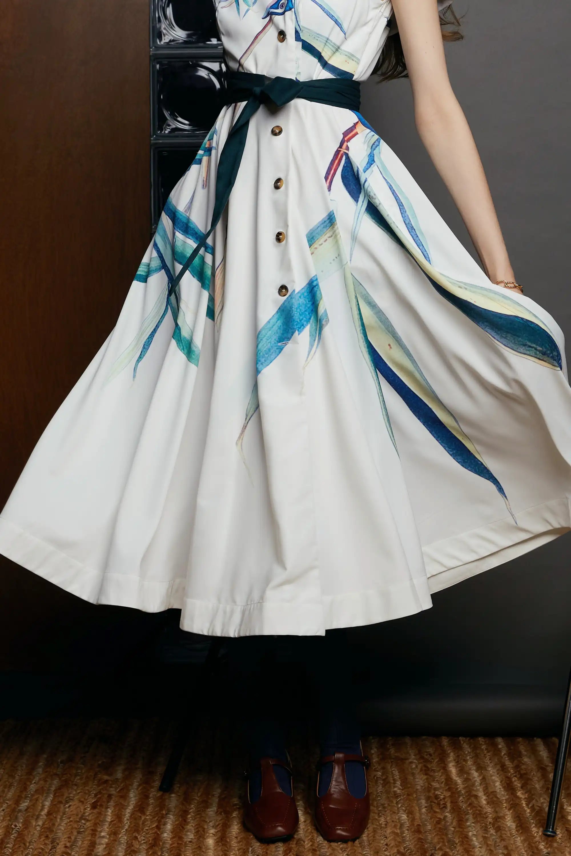 Close-up of the HAVEN Blue-headed Vireo Caplet Collar Dress showing the cream skirt with sweeping teal and cobalt brushstroke bird motifs. The dark waist tie cinches the silhouette, paired with navy socks and brown heels, captured in a studio setting.