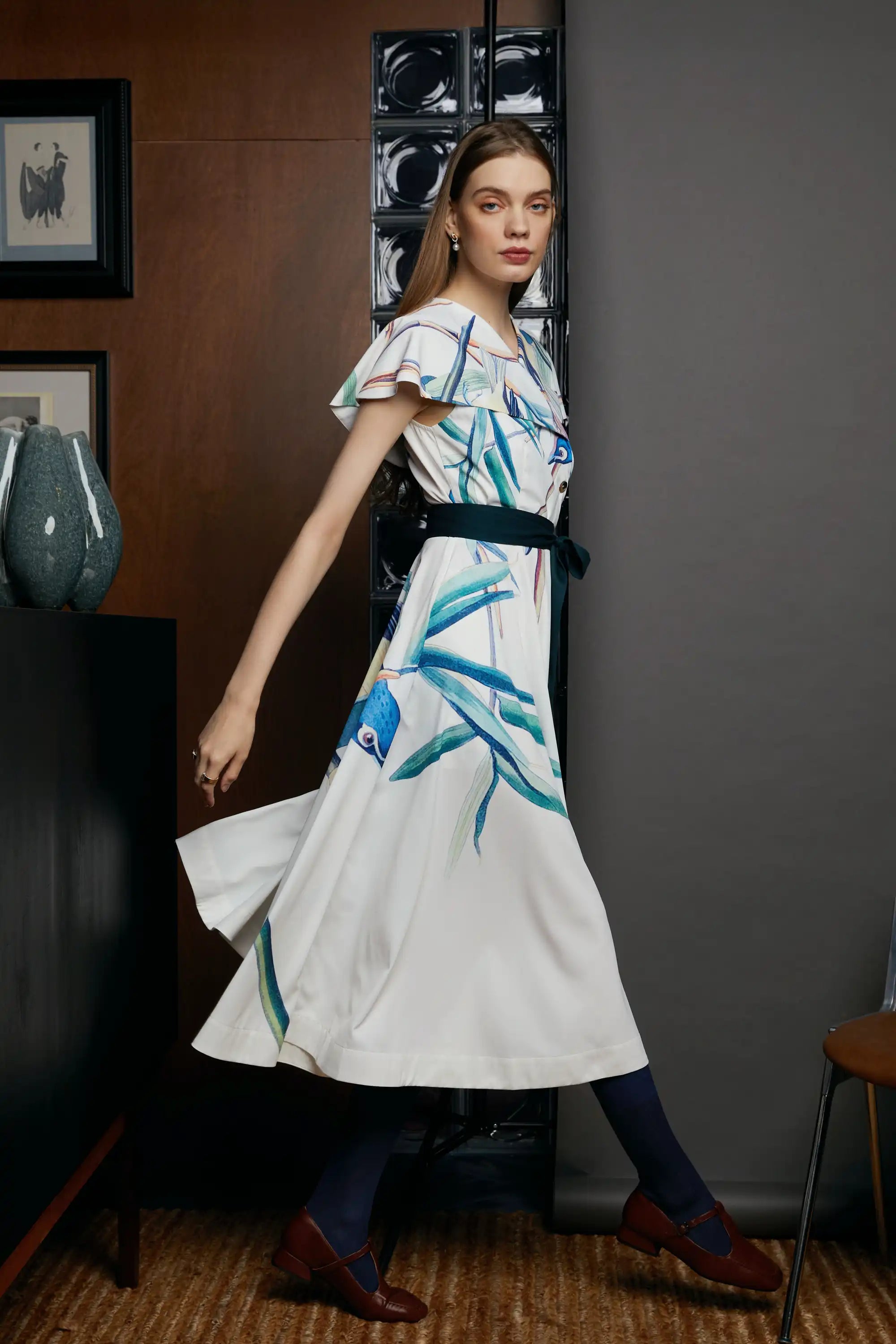 Alternate side angle of the HAVEN Blue-headed Vireo Caplet Collar Dress in cream white with teal and blue brushstroke prints. The dress is cinched at the waist with a contrasting belt and flows into a midi skirt, styled indoors with a textured grey wall and wooden panel.