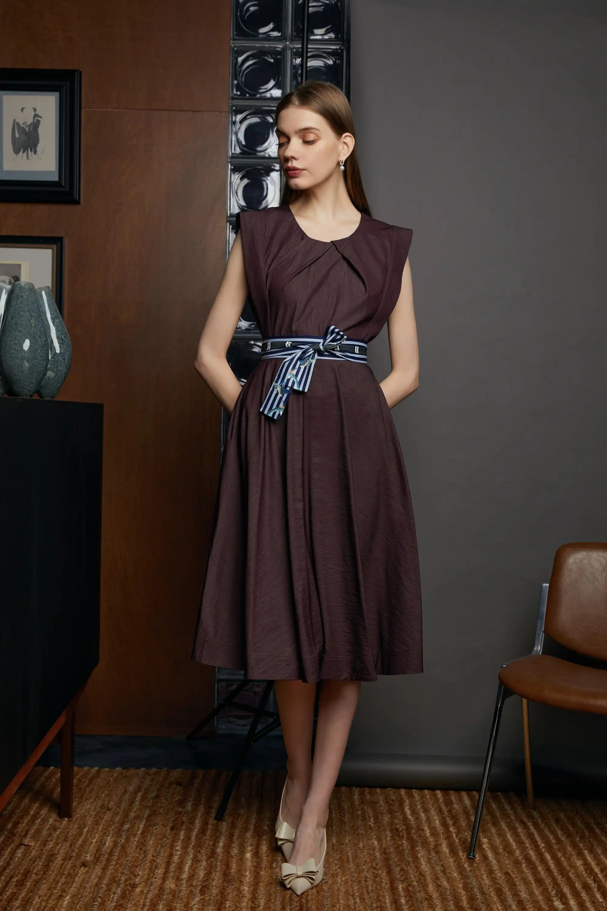 Full view of a sleeveless midi dress with square shoulders, pleated bodice, and a striped sash belt, styled with heels.