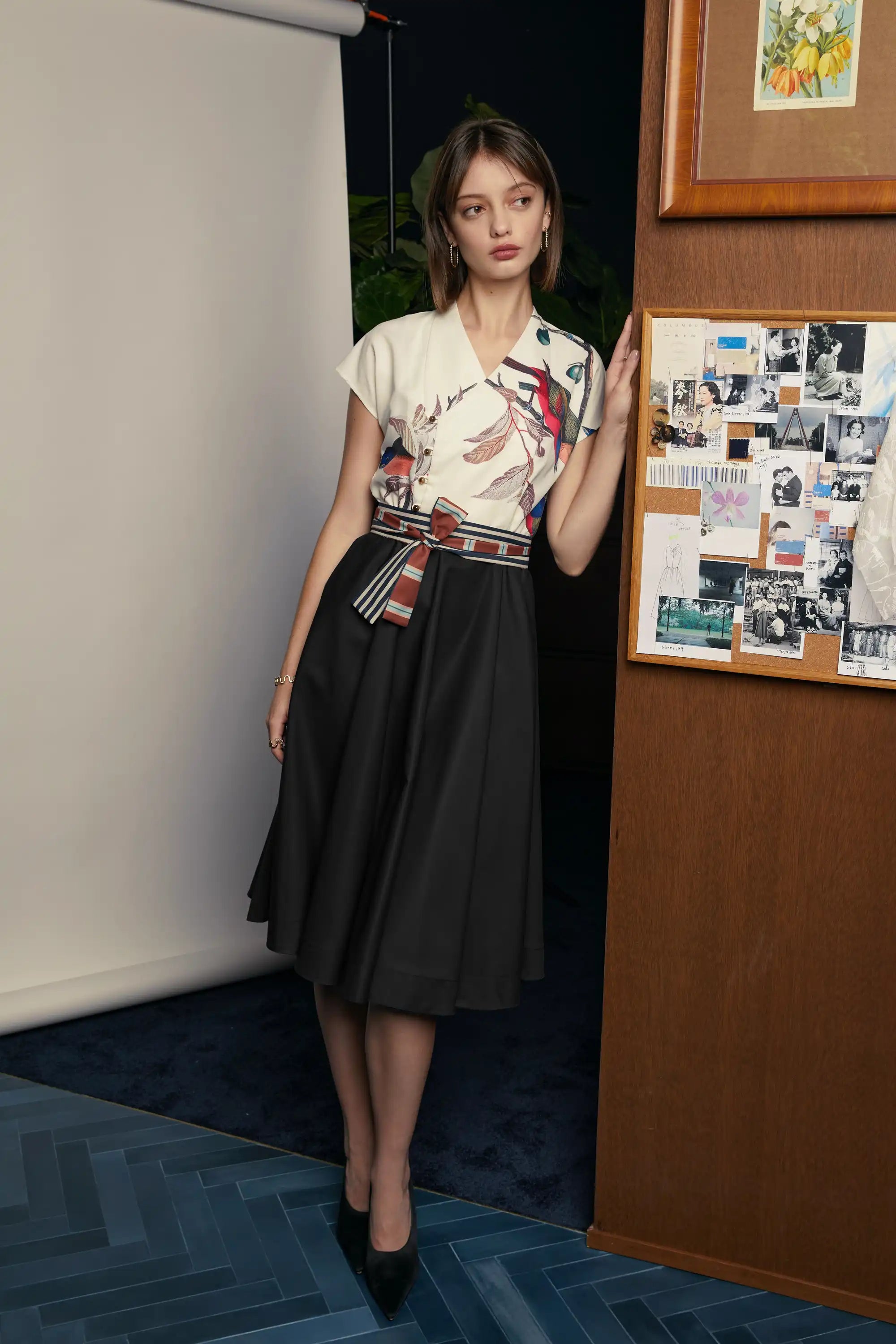 Front view of the HAVEN Surplice Fit & Flare Dress in the Painted Finch print. The dress features a cream surplice bodice adorned with bird and branch illustrations, paired with a black flared skirt. The waist is cinched with a striped sash belt that complements the print. Photographed indoors against a wooden wall and bulletin board.