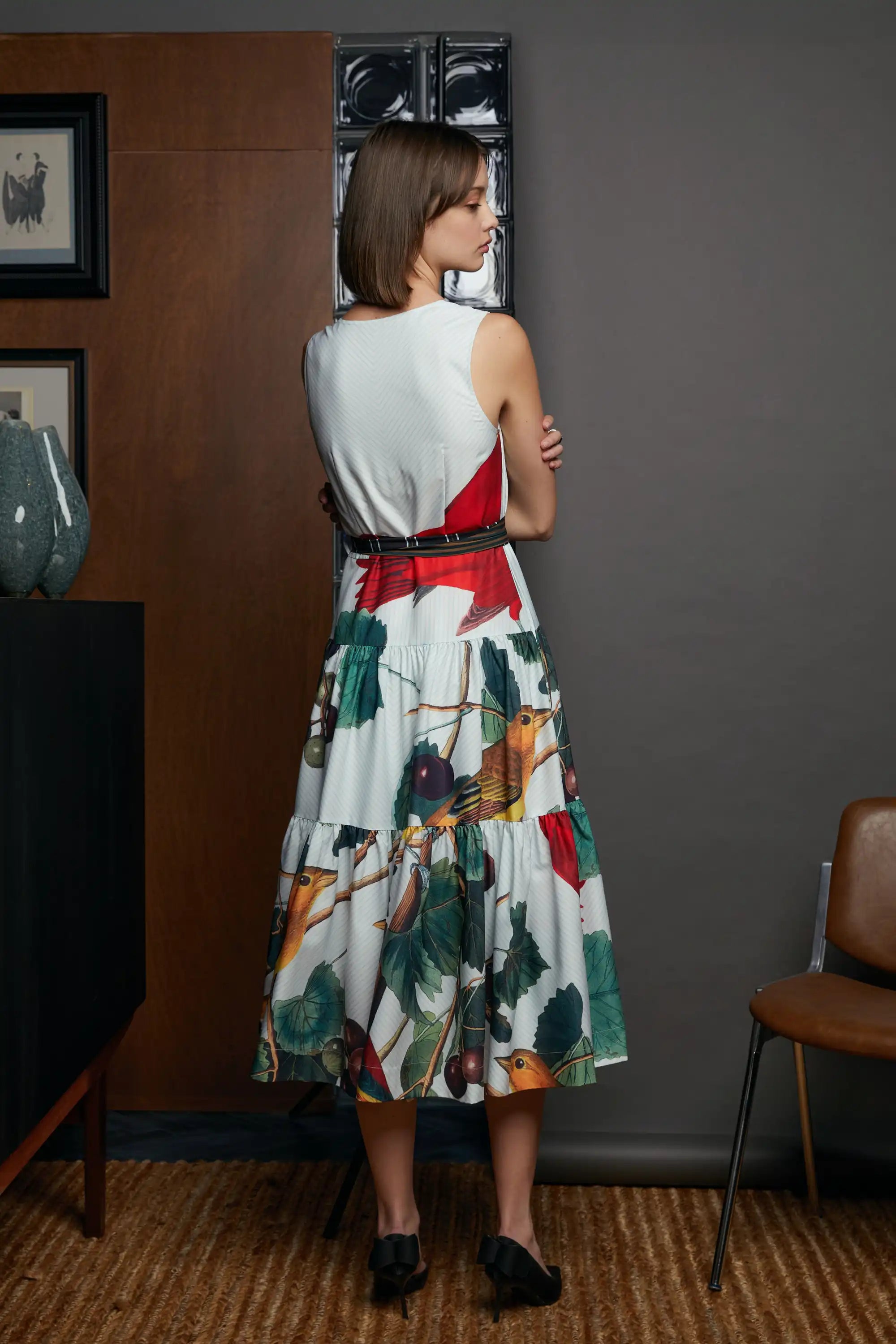Back view of the HAVEN Summer Tanager Tiered Dress, featuring the cream sleeveless bodice and a cinched waist with contrast belt, flowing into a full tiered skirt with bird, fruit and foliage prints. Styled with black heels, photographed against a grey studio wall with framed artwork and wooden floor.