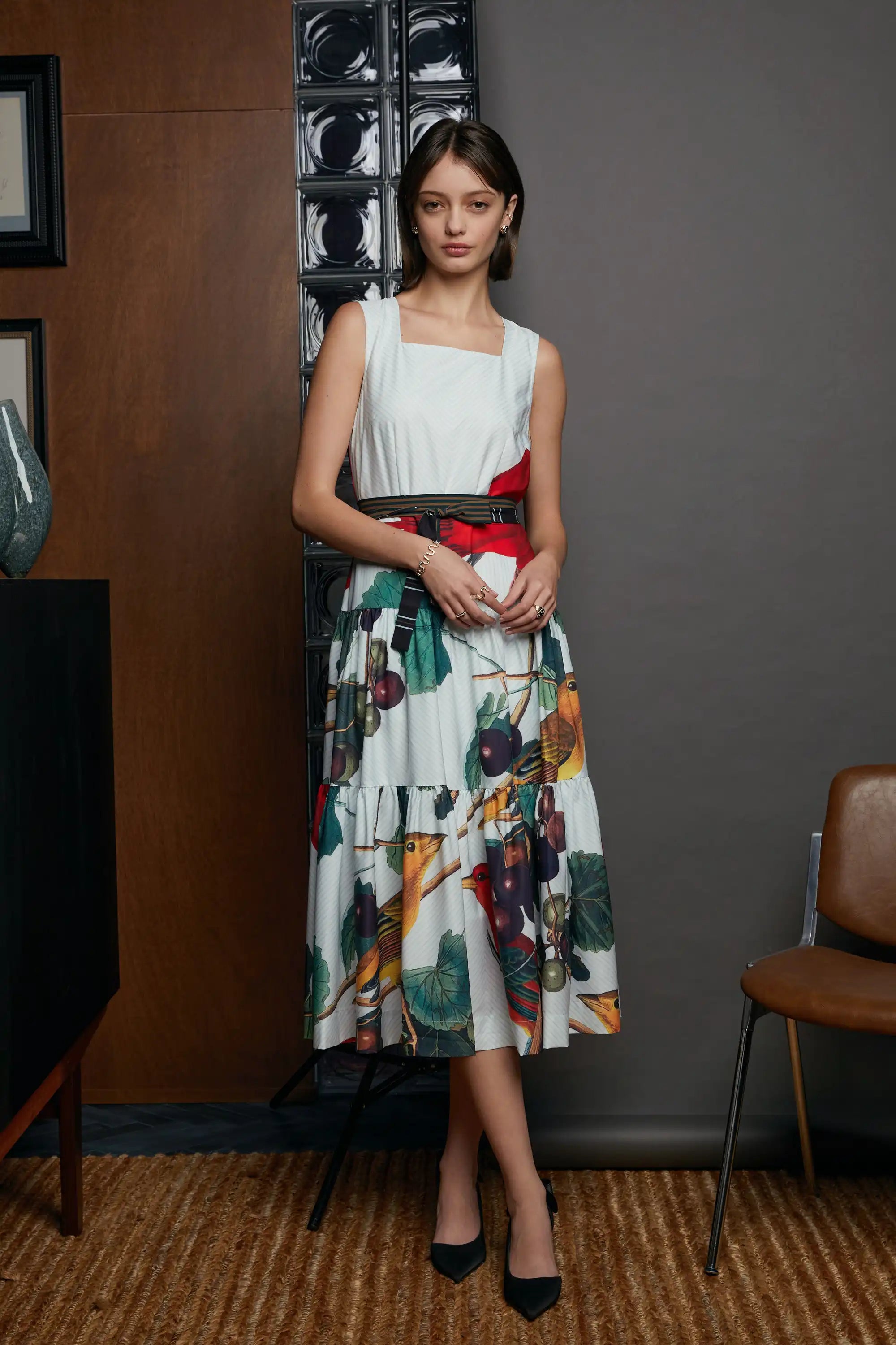 Model wearing the HAVEN Summer Tanager Tiered Dress, a cream sleeveless dress with a bold red bodice detail, cinched waist belt, and a voluminous tiered skirt featuring painterly fruit and foliage prints. Styled with black heels, photographed against a studio backdrop with wood and glass block details.