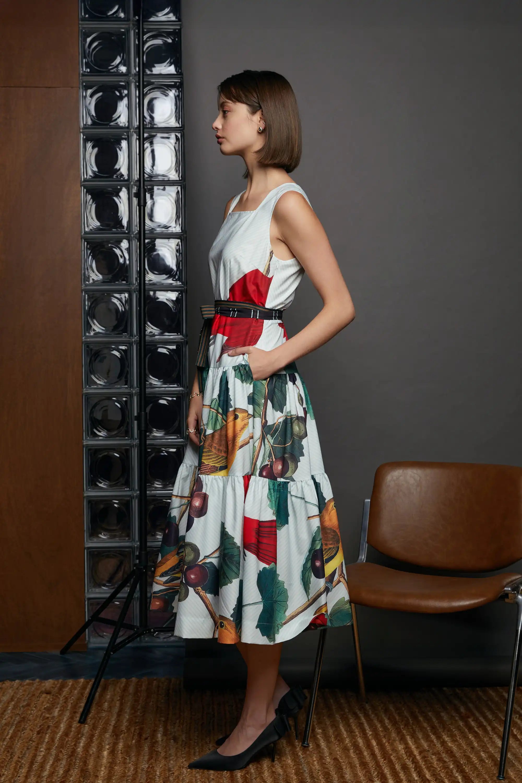 Profile view of the HAVEN Summer Tanager Tiered Dress, highlighting the red bodice accent, fitted waist with belt, and full printed tiered skirt. Model styled with black heels, photographed against a grey wall and glass block panels.