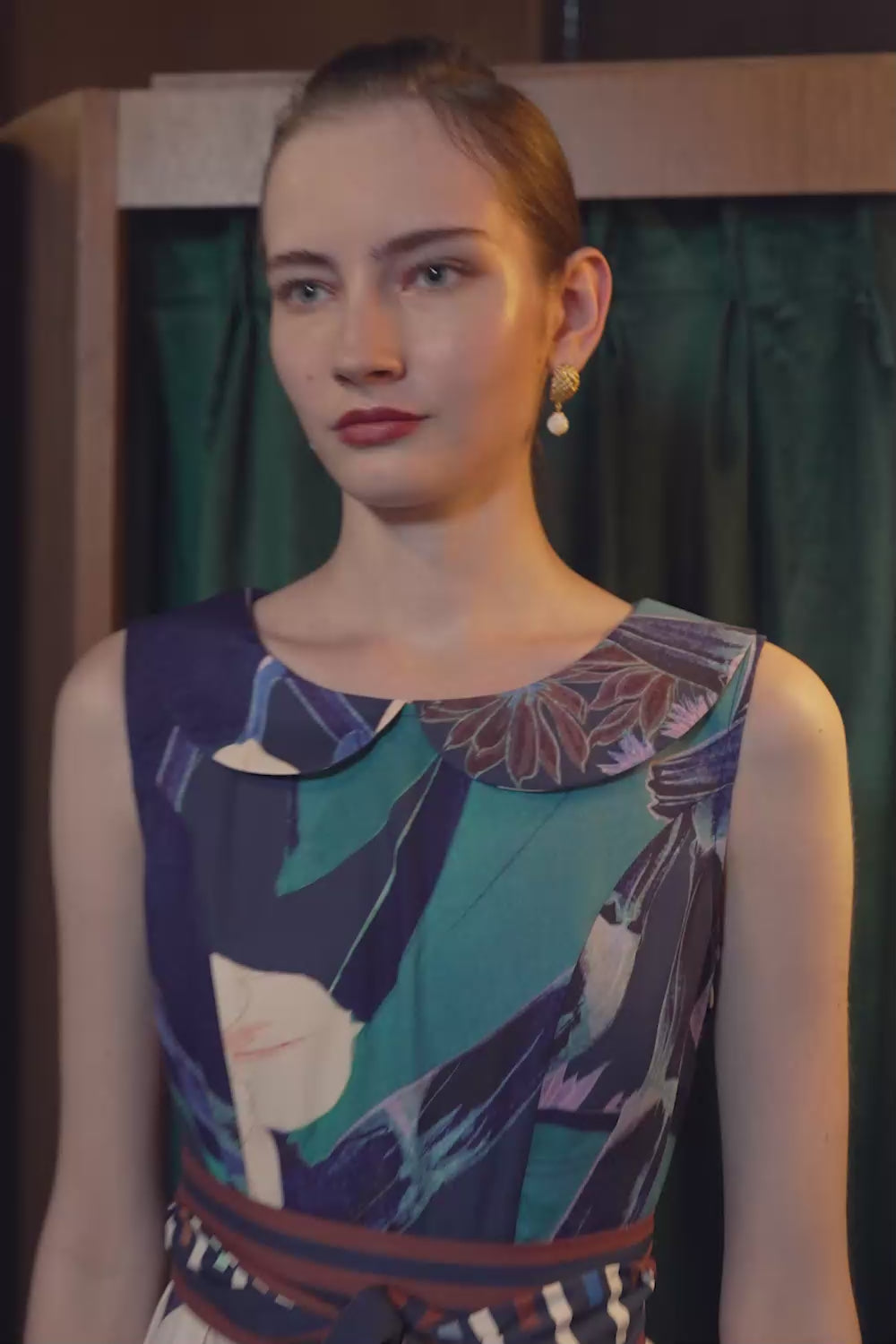 Model walking in the BEHOLD Peter Pan Fit-and-Flare Midi Dress in Dendrobium Chrysotoxum orchid print, featuring navy sleeveless bodice with Peter Pan collar, striped waistband, and cream flared skirt with orchid floral motifs, filmed indoors against a green curtain backdrop.